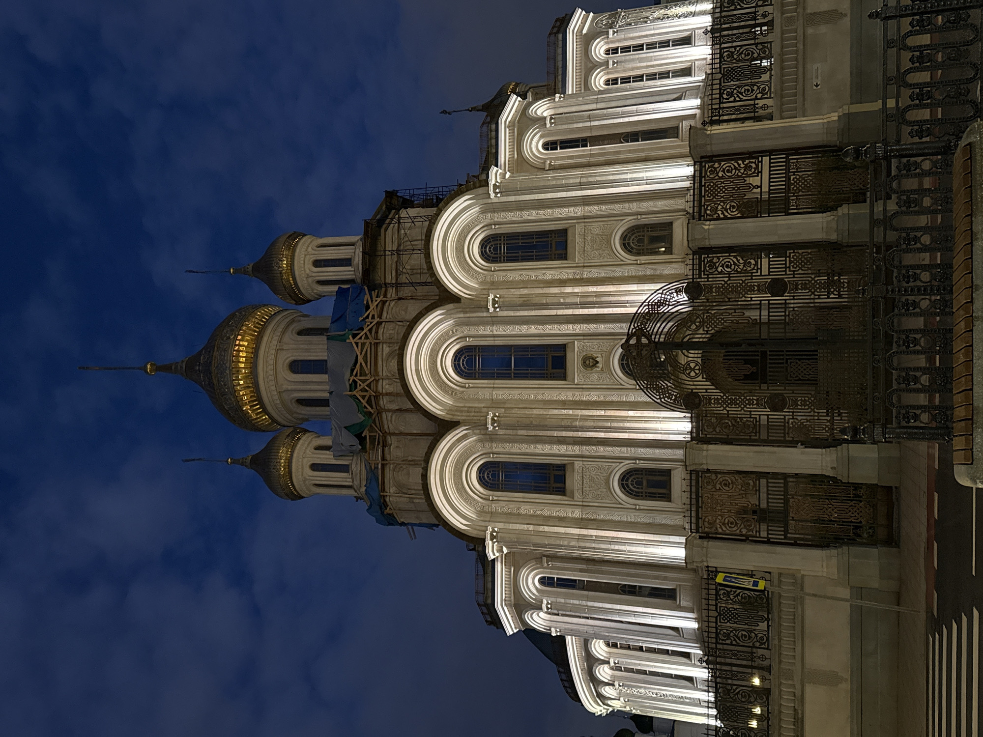 The image shows the Vysoko-Petrovsky Monastery in Moscow, Russia, at night. The monastery's ornate white facade is illuminated, highlighting its arches, windows, and decorative elements. The monastery has several golden domes topped with crosses. Some scaffolding is visible on one of the domes, indicating ongoing maintenance or restoration work. The sky is dark blue with some clouds. A wrought-iron gate is partially visible on the right side of the image, and street markings suggest the location is on a public street.