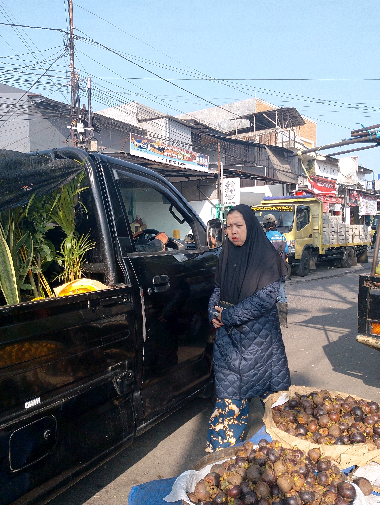 In Lembang, Indonesia, on a sunny day, a woman wearing a black hijab and a quilted dark blue coat stands beside a black pickup truck loaded with plants. She is holding a small black wallet or clutch. In front of her, on a blue mat, are two large baskets overflowing with mangosteens. Behind the pickup truck, a yellow flatbed truck is parked, carrying what appears to be stacks of concrete blocks. Further in the background, there are various buildings and a complex network of overhead electrical wires. The scene suggests a street market or a roadside vendor operation.