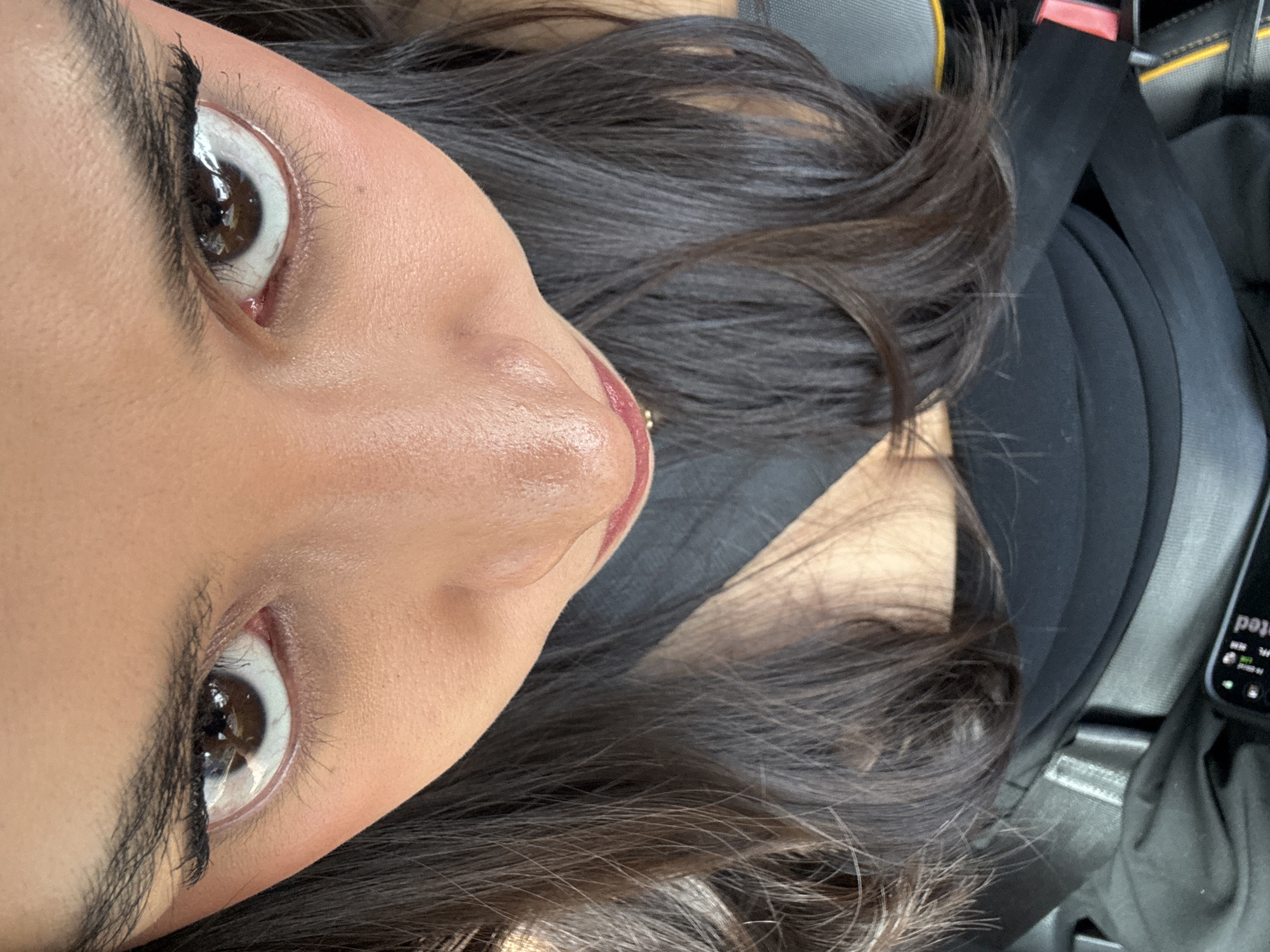 The image is a close-up, slightly angled view of a person's face, likely a selfie. The person has brown eyes and dark hair. The background shows a car interior, with a seatbelt and a mobile phone visible. Based on the phone's display, the location is Puebla, Mexico.