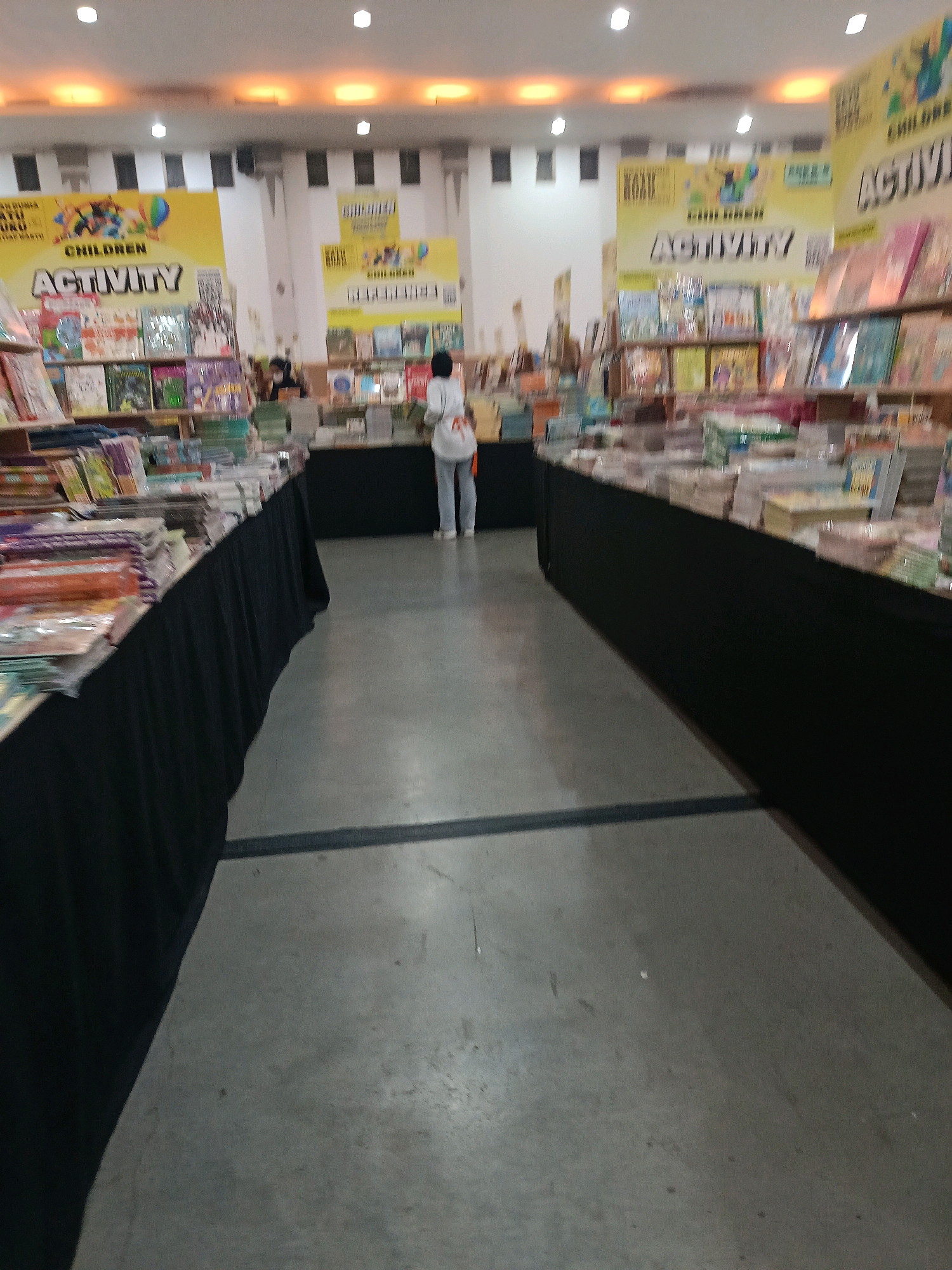 The image depicts the interior of a bookstore, likely at a book fair, as indicated by the displays and the presence of books. The location is identified as Banguntapan, Indonesia. In the foreground, a long table draped with a black cloth displays numerous books. To the right, a person wearing a white shirt and light-colored pants is walking away from the camera, carrying a white bag. Further back, a second person in a black hijab is visible behind a counter, possibly attending to customers. The background features yellow banners with text and illustrations, advertising "CHILDREN ACTIVITY." The lighting suggests an indoor setting with overhead lights visible on the ceiling. The floor is a gray concrete surface with black lines.