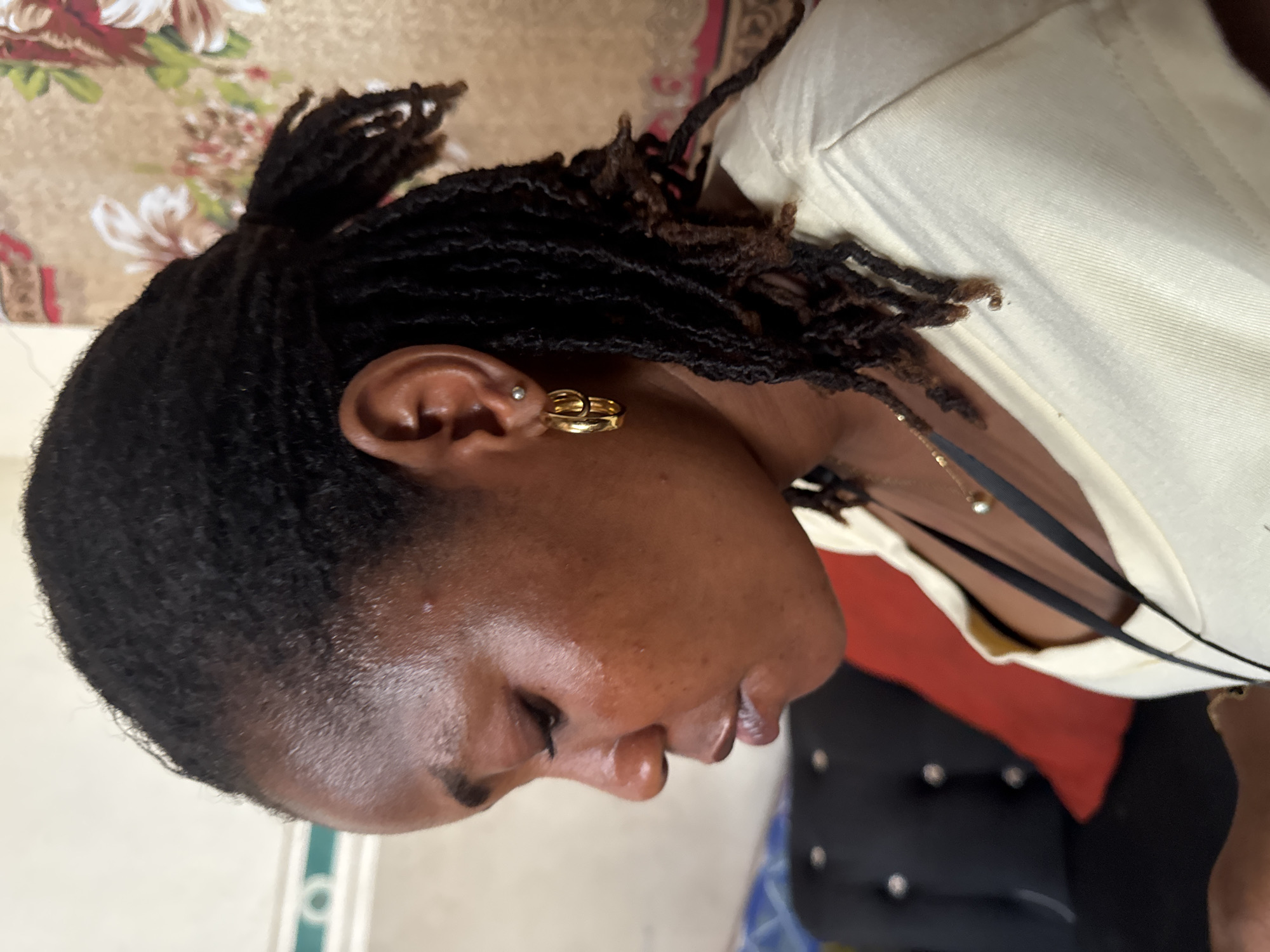 A close-up, profile shot captures a Black woman with locs pulled into a partial updo. She wears gold earrings: one a small stud in the upper ear, the other a more elaborate design on the lobe. Her skin appears dewy. A pale yellow shirt is visible, along with thin black straps. The background is a blend of patterns, including floral designs, and glimpses of furniture. There is no evidence to place this scene in Badagry, Nigeria.