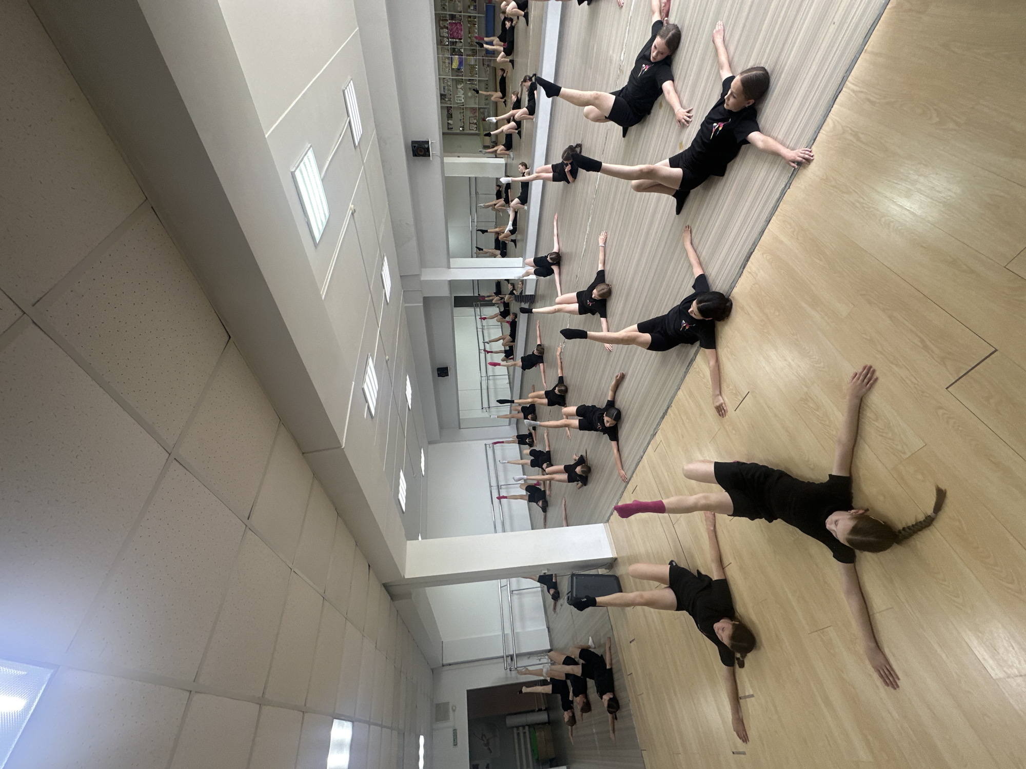 This media file captures a practice session or class for a group of young people, primarily girls, in an indoor studio. The subjects are positioned on a light-colored wooden floor, lying on their sides with one leg extended upwards and both arms stretched out, forming a 'T' shape with their bodies. This synchronized pose suggests a disciplined exercise, warm-up, or dance routine.
The participants are uniformly dressed in black short-sleeved t-shirts and black shorts. A small, red or pinkish stylized logo or emblem is visible on the chest of their t-shirts, though the specific design or text is too indistinct to be read. Hair is mostly tied back, with some individuals sporting braids. One detail includes a participant wearing a single pink sock on their extended foot.
The setting is a brightly lit studio or dance hall. A prominent feature of the room is a large mirrored wall that reflects the participants and the interior space, creating the appearance of multiple rows of individuals performing the same pose. The ceiling is fitted with rectangular fluorescent light fixtures and acoustic ceiling tiles. The overall ambiance is one of focused activity within a well-equipped practice environment. There are no visible cues to determine the time of day, given the artificial lighting. The location context is Votkinsk, Russia.