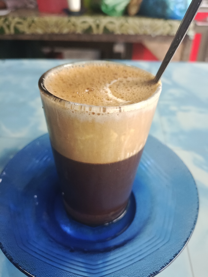 A glass of layered coffee rests on a blue saucer. The bottom layer of the drink is dark brown, topped by a lighter brown layer, and finished with a foamy tan layer at the top. A black straw or stirrer protrudes from the drink. The saucer is a clear, textured blue with a simple geometric pattern. The background is slightly blurred, showing a table with other objects.