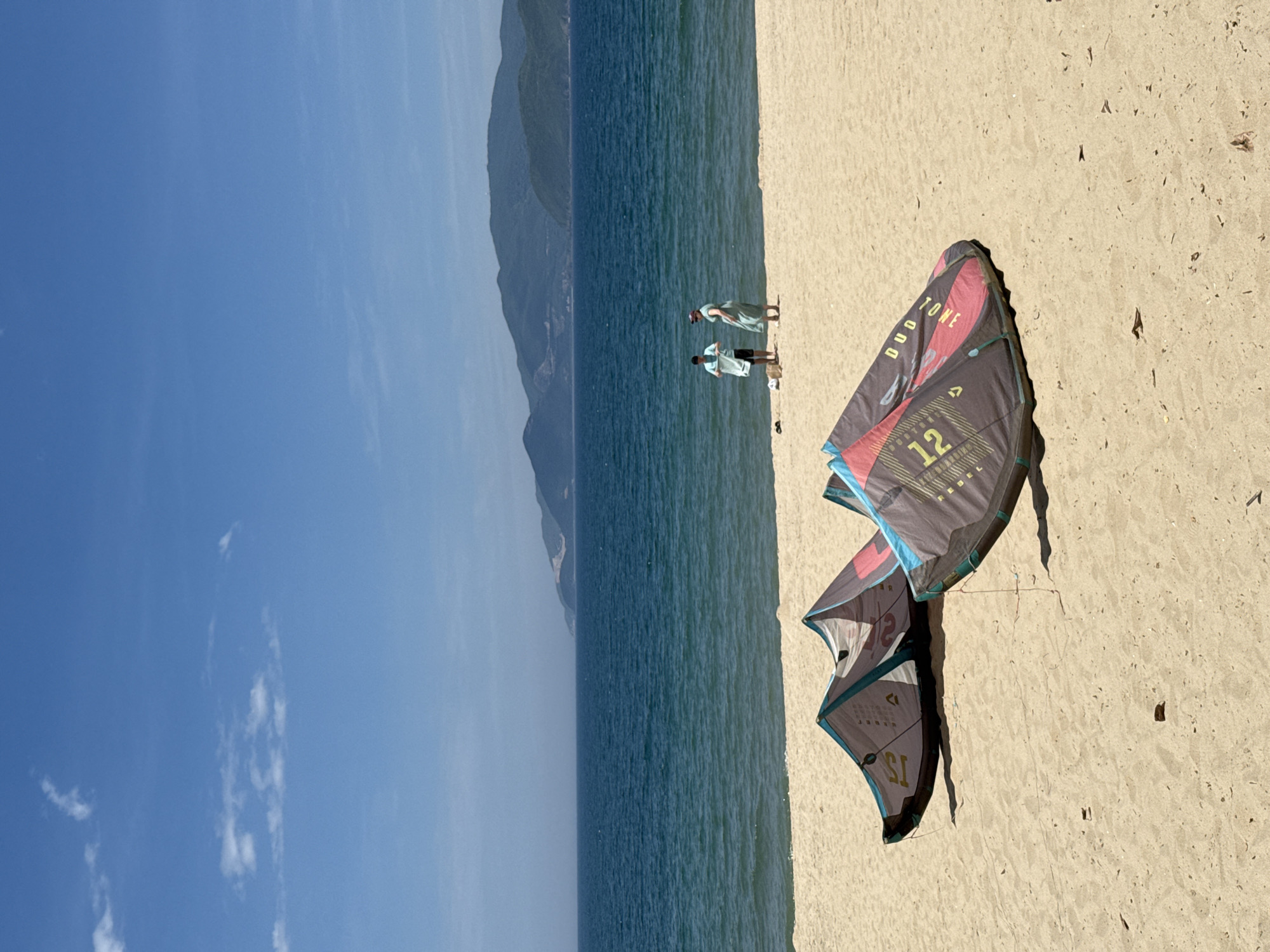 This vertical, eye-level, outdoor, full-shot photograph captures a serene beach scene in Nha Trang, Vietnam, under a clear blue sky dotted with sparse white clouds. The foreground is dominated by the sandy beach, where two kiteboarding kites are laid out. The kite on the left is predominantly grey, white, and teal, featuring faded text and a number '1'. The kite on the right is larger and more vibrantly colored, with sections of black, grey, red, and teal, displaying the text 'DUO TONE' and the number '12'.
In the middle ground, the turquoise ocean stretches to the horizon, meeting a range of lush, green, mountainous islands in the distance. Two individuals are present on the sand near the water's edge. The person on the left, wearing a light blue t-shirt and dark shorts, is facing away from the camera and appears to be holding a white item, possibly a towel. The person on the right, dressed in a light green dress and a dark head covering, is facing the other individual.
The environment is tranquil, with the calm sea and vast sky suggesting a peaceful day. The presence of kiteboarding equipment indicates a location popular for water sports. The overall atmosphere is bright and sunny, typical of a tropical coastal setting.