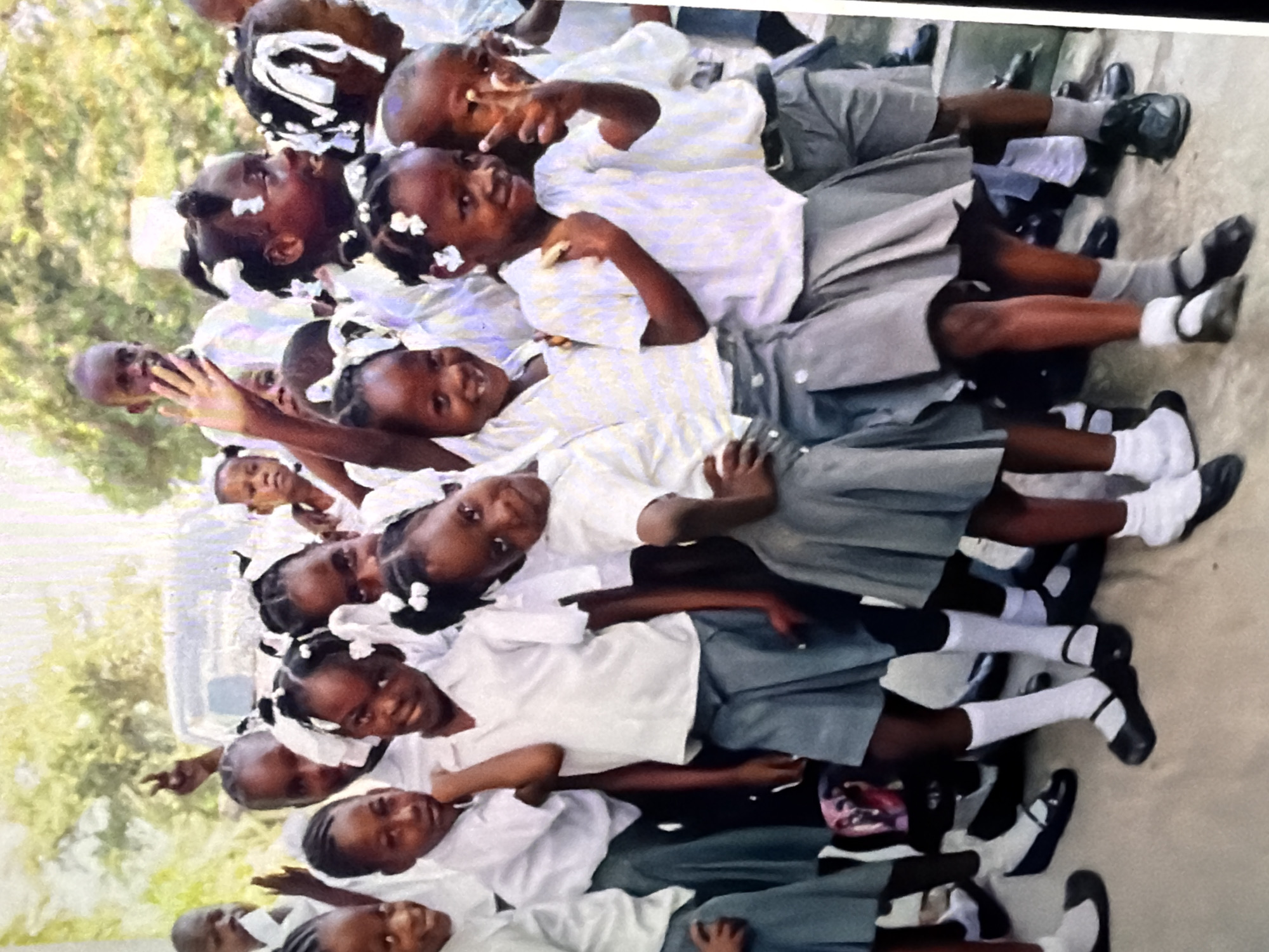 A group of young African school children, dressed in white tops and grey skirts, stand together outdoors. Some of the girls have white bows in their hair. They are posing for the camera, with some waving and others making peace signs. The background is blurred, but appears to be a sunny day with trees and a vehicle visible.