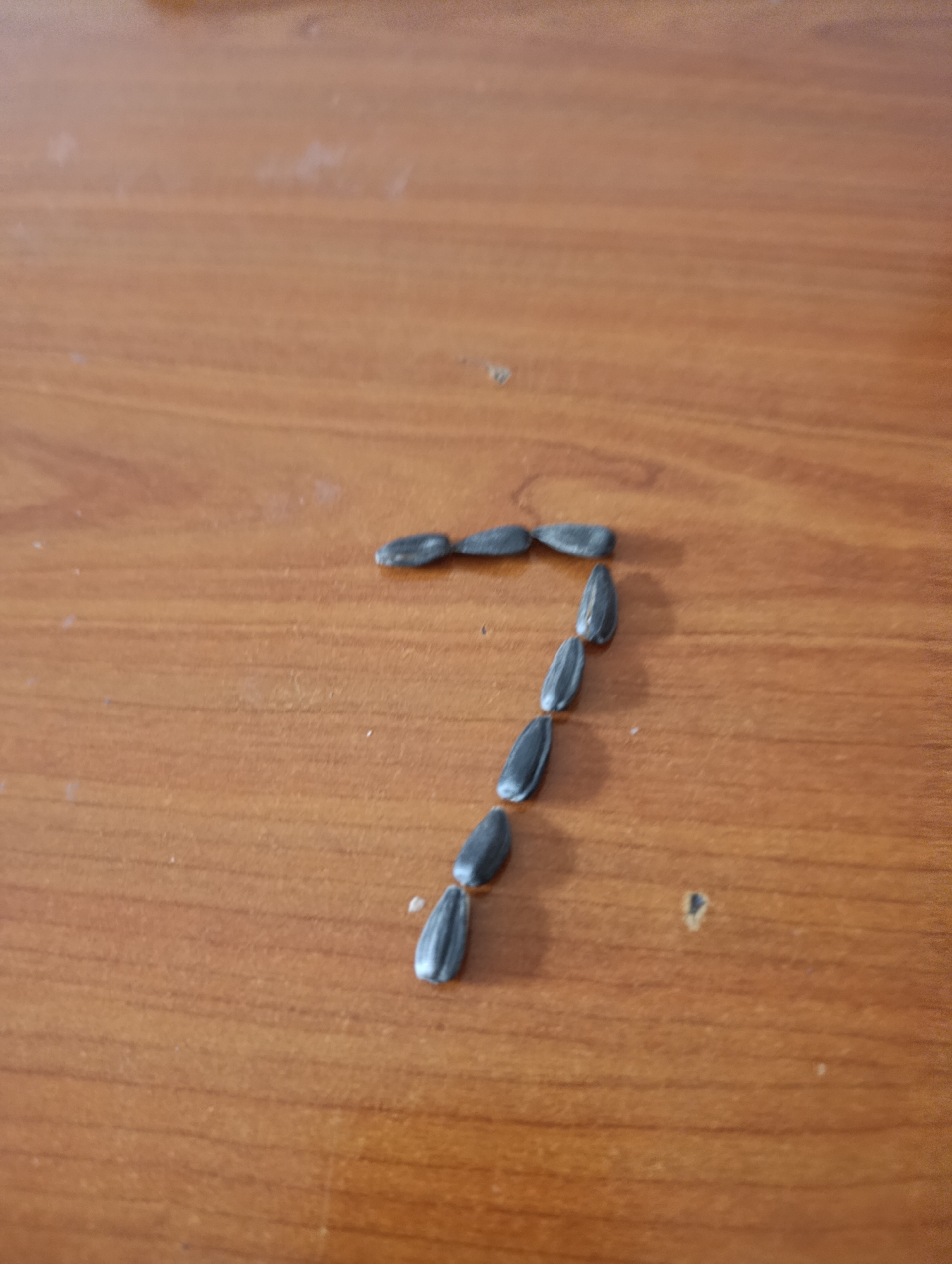 The image shows the number 7 formed by seven sunflower seeds arranged on a brown wooden surface. The seeds are oriented to form the digit, with three seeds horizontally across the top and four seeds descending diagonally downwards. The background is a smooth, slightly blurred brown wooden surface. The location is unknown.