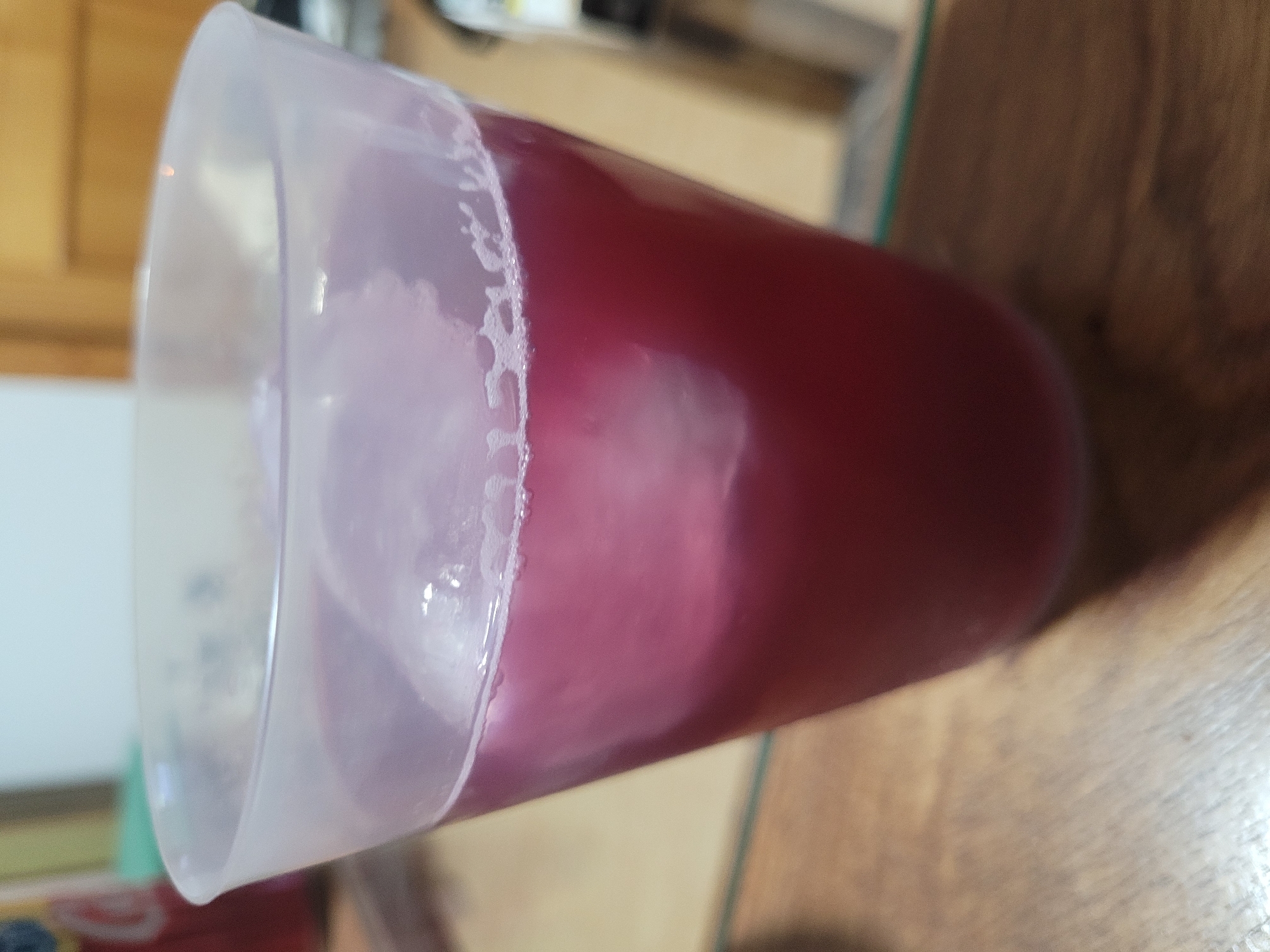A close-up, slightly angled shot shows a clear plastic cup filled with a reddish-purple liquid and ice cubes. The cup has a frosted texture and a slightly flared rim. The liquid has a foamy layer near the top. The cup is placed on a wooden surface. In the background, out of focus, a red object with some text and a green object are visible. The environment appears to be indoors, possibly a kitchen or dining area, with wooden doors and walls in the background. The image does not provide any specific visual cues or context that would allow for the determination of its location in Els Campellos, Spain.