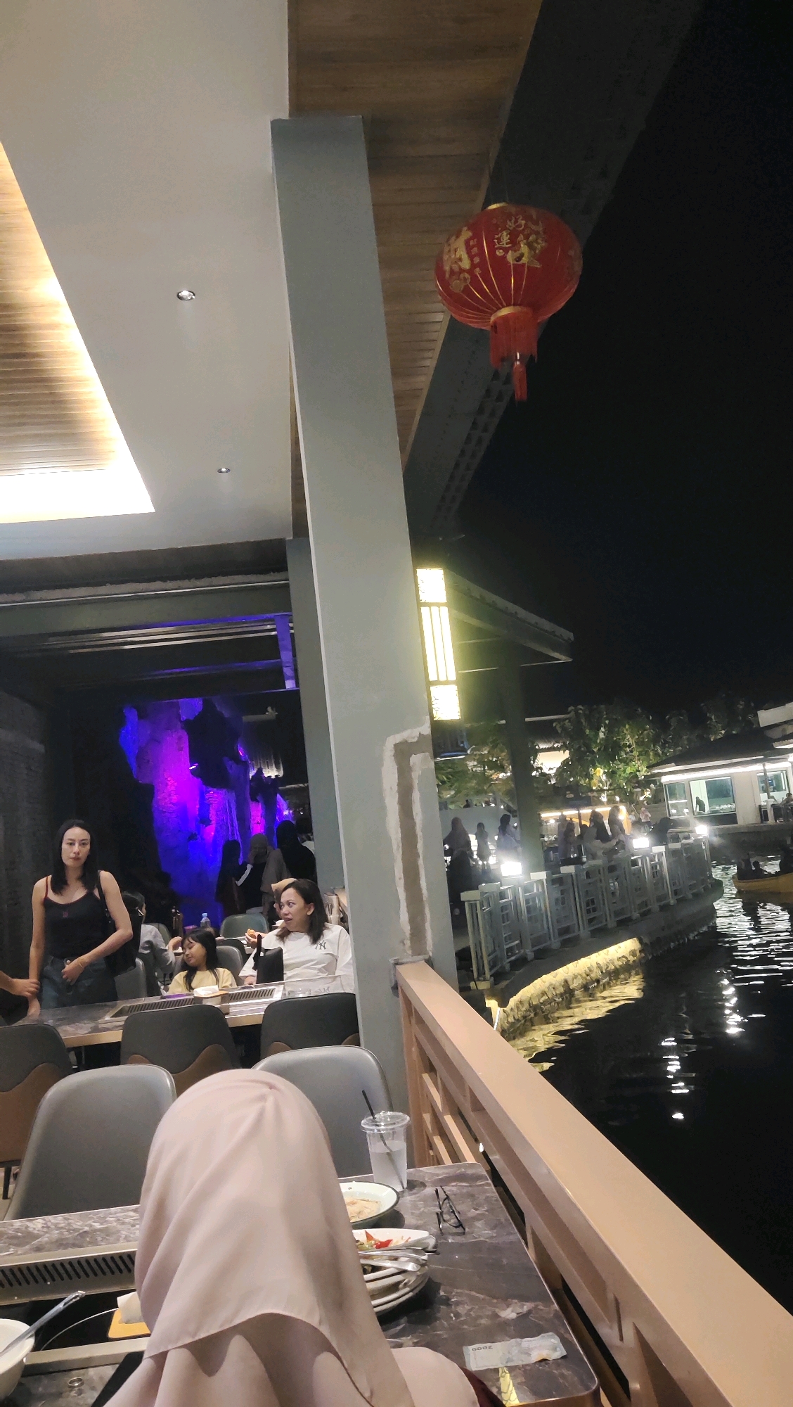 The image depicts an outdoor dining area at night, likely a restaurant with a waterfront setting. In the foreground, a person wearing a light pink hijab is seated at a table, with their back to the camera. The table is laden with dishes, cutlery, and a glass of water. To the left, a woman in a black tank top stands, and a young girl in a yellow top is seated. Further back, a woman in a white shirt is seated and looking to her left. In the background, more diners are visible at tables lining a waterway. The restaurant appears to have an Asian theme, suggested by a red lantern hanging from above and illuminated rock formations with purple lighting. The overall ambiance is casual and social. The location is specified as Pandak, Indonesia.