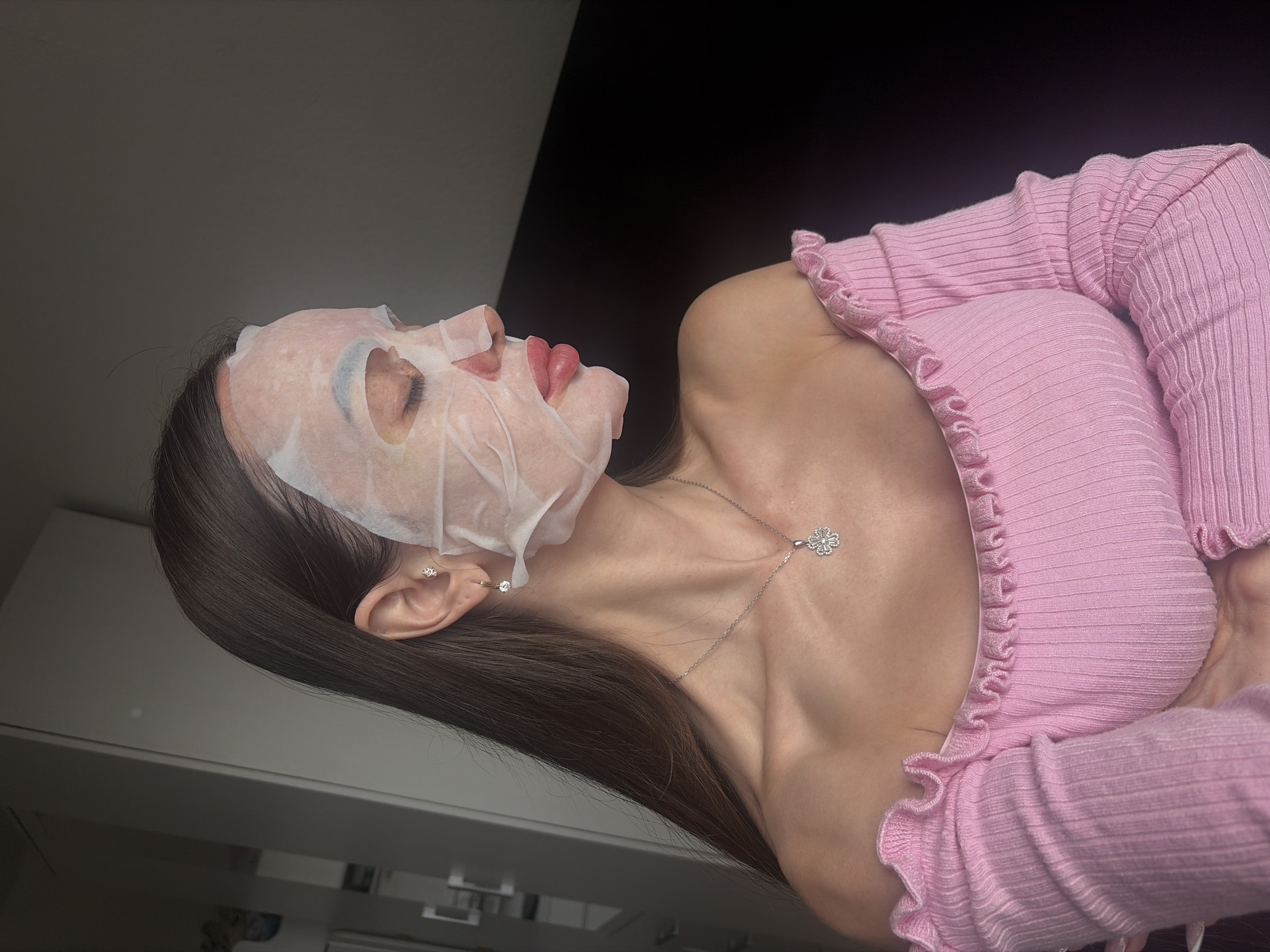 A woman wearing a pink off-the-shoulder ribbed sweater and a silver necklace with a flower pendant is lying on her back, wearing a white sheet mask on her face. She has long, dark hair and wears earrings in her left ear. The background consists of a black wall on the upper right and a light-colored wall with furniture on the lower left. The image does not contain sufficient information to determine its location in Saint Petersburg, Russia.