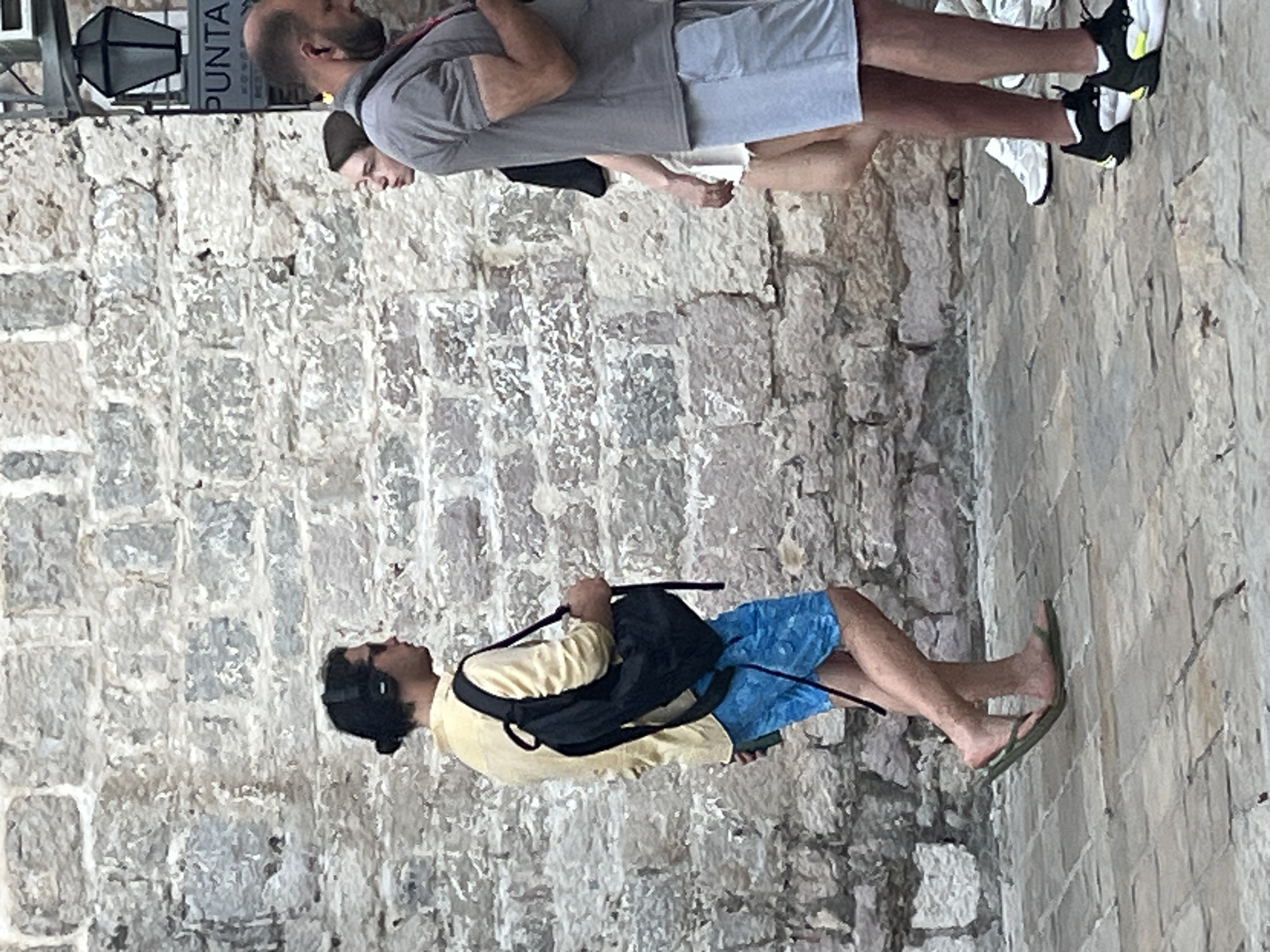 In Budva, Montenegro, on a cobblestone street with a rough, stone-block wall as a backdrop, three individuals are depicted in motion. The foreground features a man from the waist down, wearing dark athletic shoes with yellow accents, and light blue shorts, walking right. Behind him and to his left, a man in yellow short-sleeved shirt and blue patterned shorts, wearing headphones and carrying a black backpack, walks right. To the right of the man in the yellow shirt, a woman in a white skirt and a grey-shirted man are walking away from the viewer, towards the right of the frame. A decorative street lamp is visible on the wall, with a sign reading "PUNTA" below it. The scene suggests a public, outdoor urban environment.