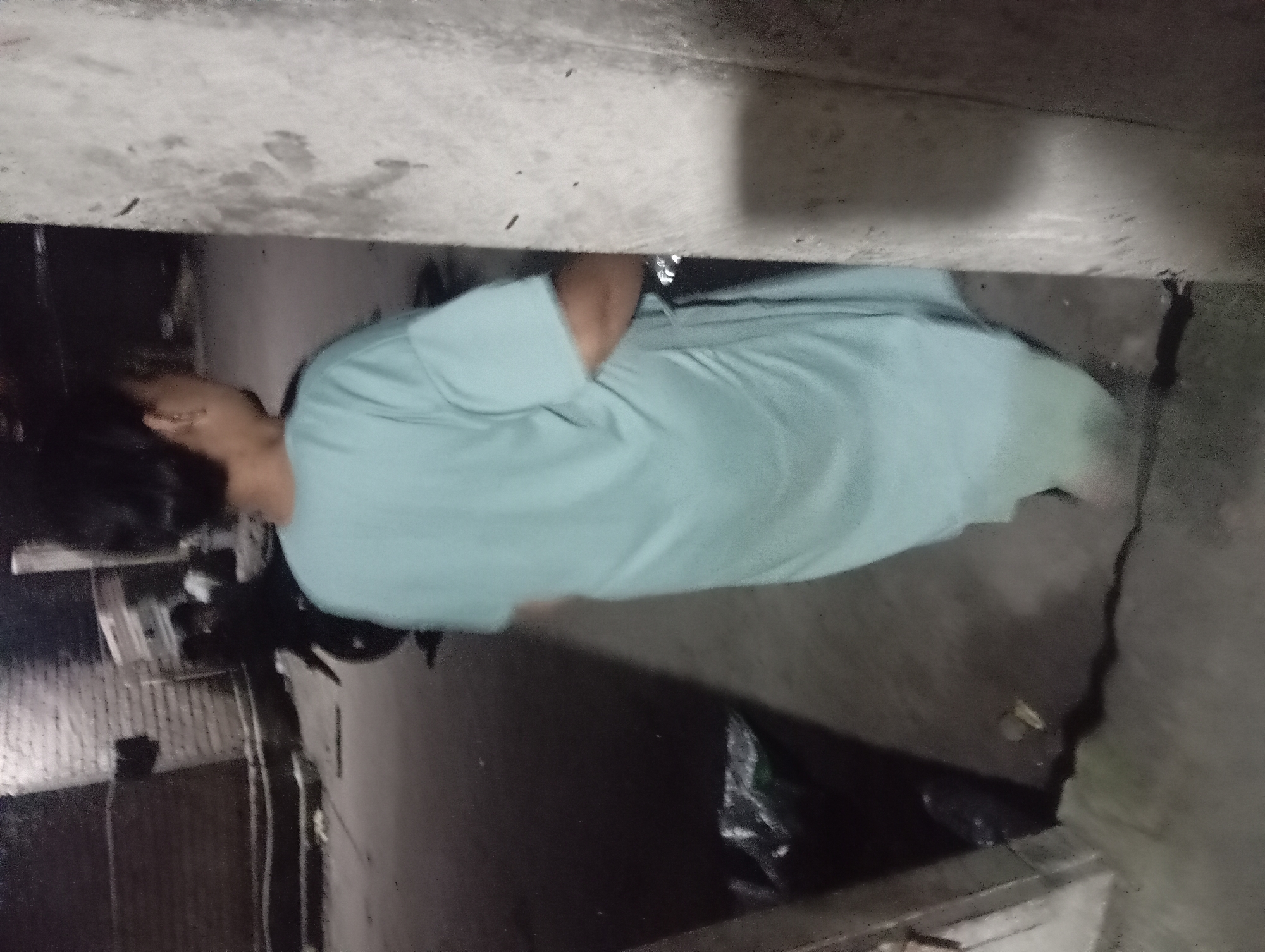 A person wearing a light blue oversized t-shirt stands in a dimly lit outdoor area, with their back to the camera. The environment appears to be an alleyway or narrow street in Rejosari, Indonesia, characterized by a rough concrete wall on the right, a brick wall in the background, and a dark, possibly wet, ground surface. A motorcycle is visible in the background, with another person sitting on it. There are some black plastic bags on the ground to the left of the frame. The overall lighting is low, suggesting it is nighttime or a very shaded area. The image is grainy, indicating it was likely taken with a low-quality camera or in poor lighting conditions.