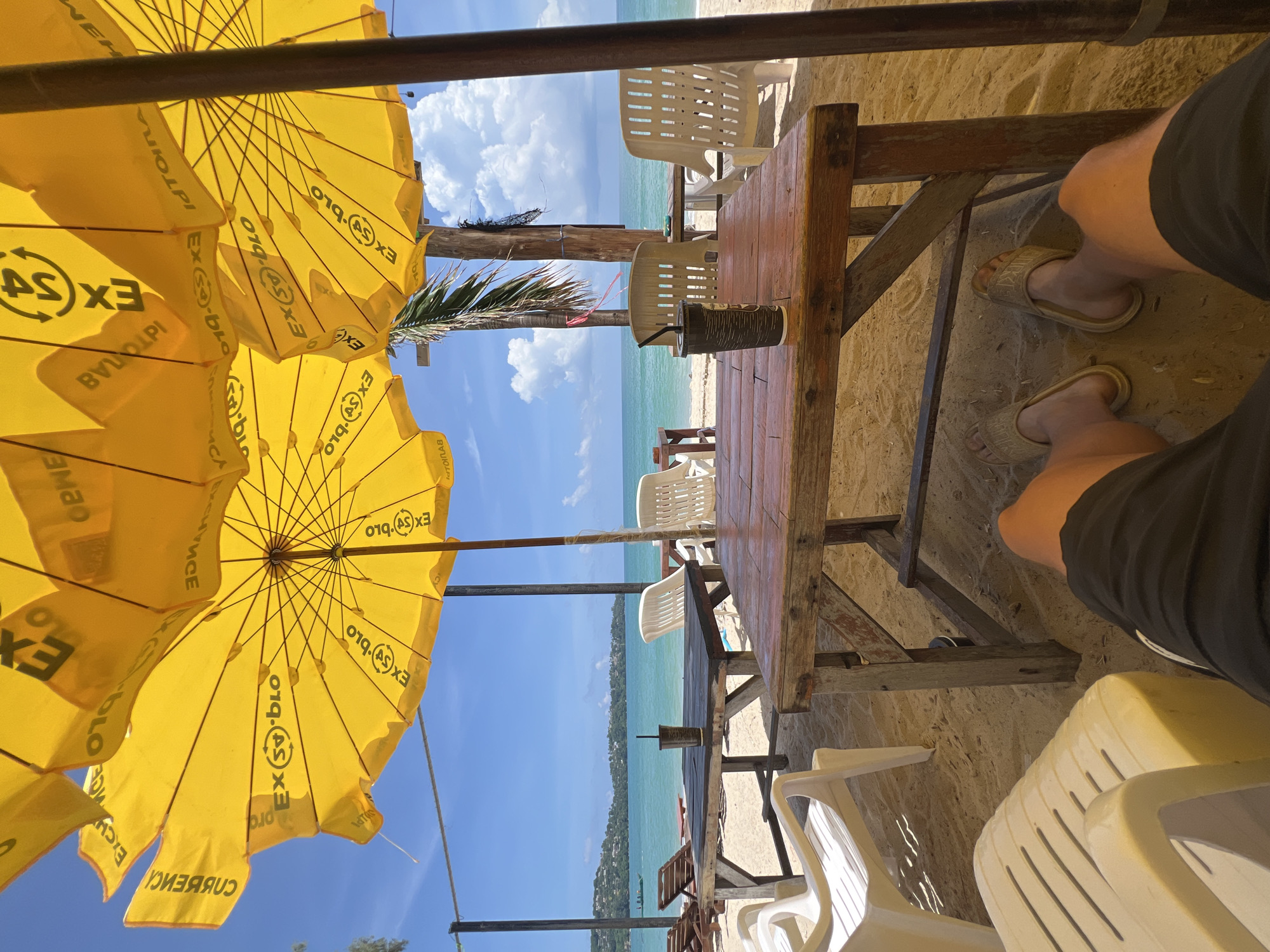 A first-person perspective shot captures a beachside scene under large yellow umbrellas. In the foreground, a person's legs and feet, wearing black shorts and light-colored sandals, are visible, resting on the sandy ground. In front of them is a wooden table with a dark beverage in a cup and a straw. Behind the table, white plastic chairs are arranged, and a few lounge chairs are visible further back. The beach overlooks a bright blue ocean with a distant, tree-lined coastline. The sky is clear blue with fluffy white clouds. The yellow umbrellas have a repeating logo with the letters "EX" and a circular symbol.