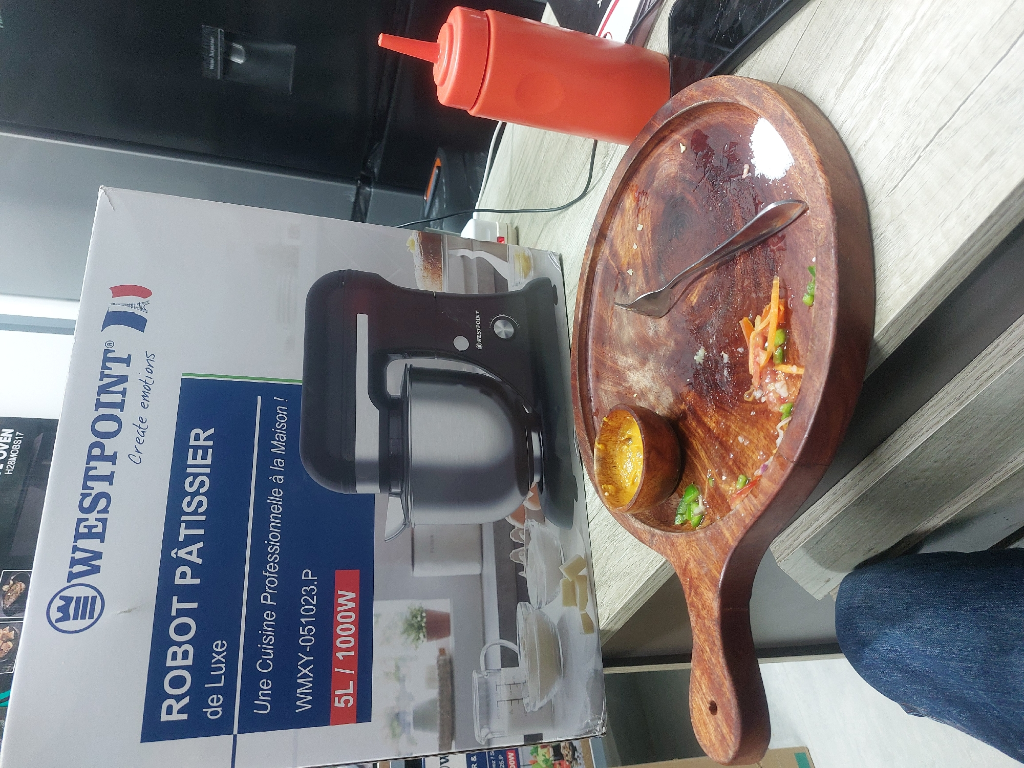 The image depicts a countertop scene. Prominently displayed is the packaging for a "WESTPOINT Robot Pâtissier de Luxe" stand mixer, with details like "5L/1000W" and model number "WMXY-051023.P" visible. Next to the packaging is an orange squeeze bottle. A wooden serving platter with food remnants and a small bowl containing what appears to be sauce sit next to the mixer box. A fork rests on the platter. A person wearing blue jeans is partially visible at the edge of the image. The location is inferred to be in Dar es Salaam, Tanzania, based on the language used on the mixer box.