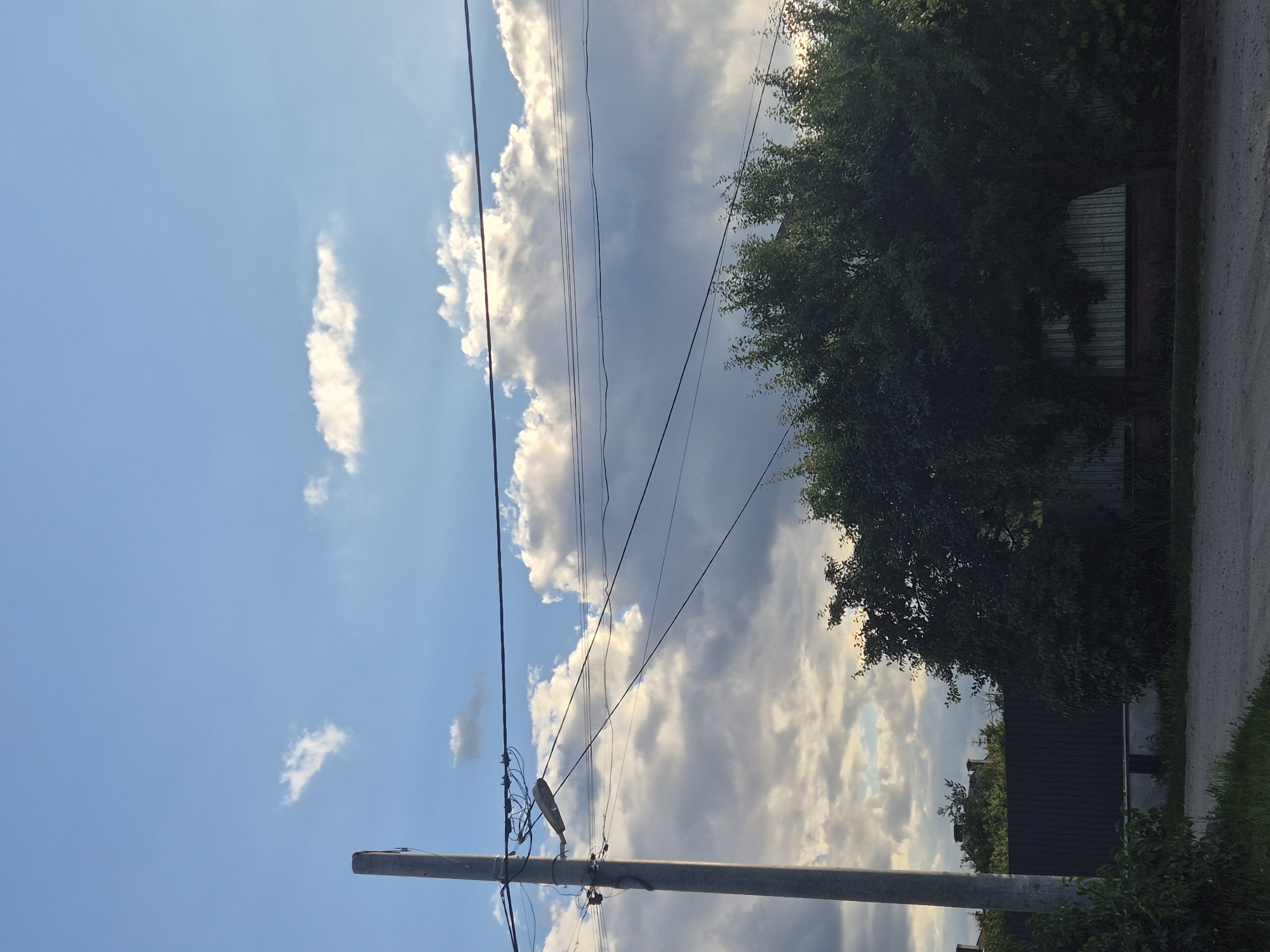 A vertical, gray utility pole stands on the left side of the image, equipped with a street lamp and a complex network of overhead wires. These wires crisscross horizontally through the frame. The sky above is a mix of bright blue and large, white, cumulus clouds, some of which are illuminated by sunlight, casting soft shadows. In the middle ground, a dense cluster of green trees obscures a dark, corrugated metal fence and a glimpse of a building. The foreground features a gravel road with some green vegetation on the left edge. The scene appears to be set in a rural or suburban area, likely Sozonivka, Ukraine, given the presence of utility infrastructure and the natural vegetation. There are no people visible in the image.