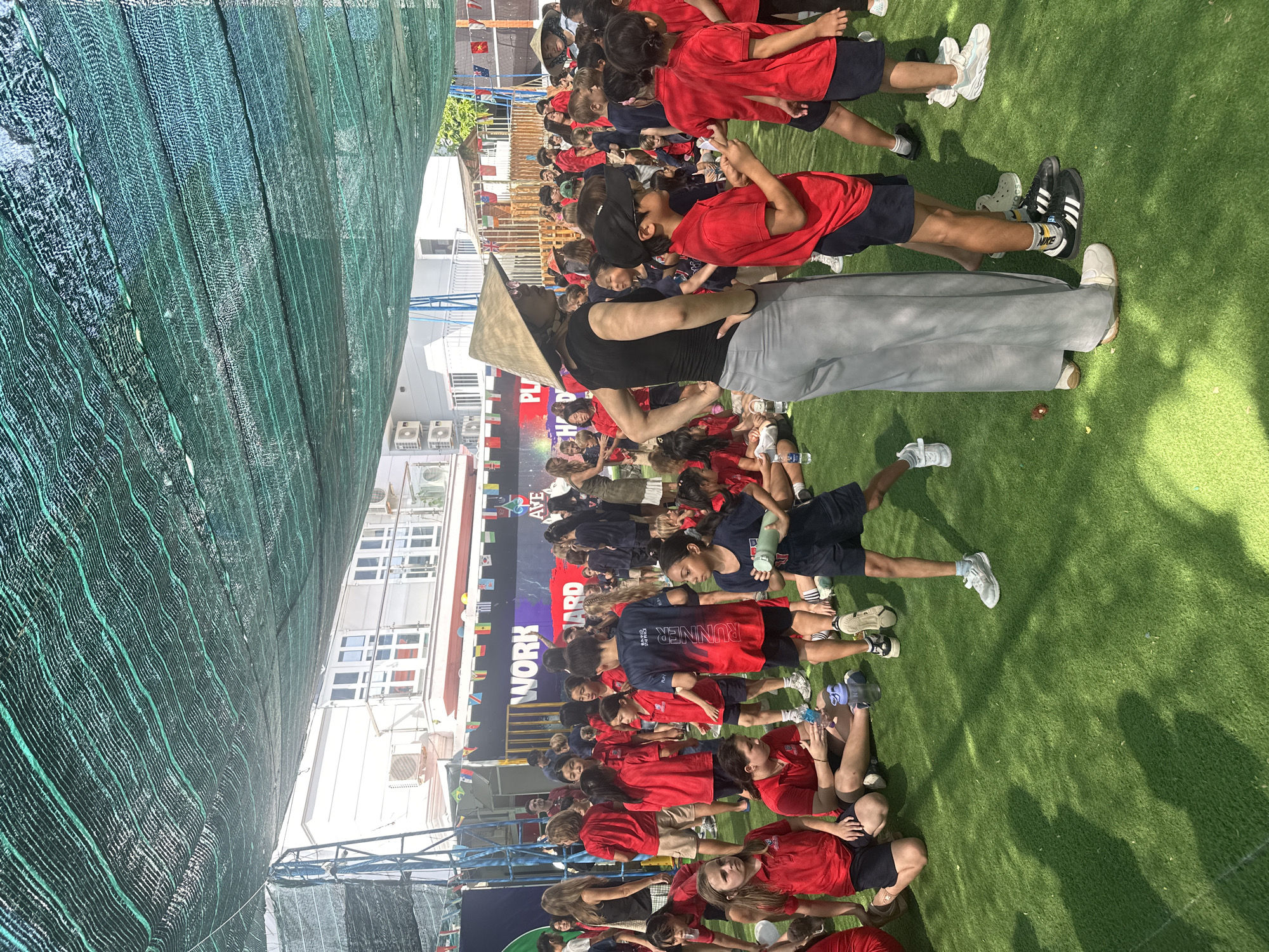The image depicts an outdoor event, likely at a school, with a large gathering of children and some adults on a bright, sunny day. A green mesh netting is stretched overhead, providing shade. The ground is covered with artificial green turf. In the background, there's a white building with multiple windows, some of which appear to be balconies. A banner with the words "WORK" and "HARD" is visible on a purple backdrop. Colorful flags are strung across the scene.
The majority of the people are children, dressed in red shirts and dark shorts, suggesting a school uniform. Some adults are also present, including one wearing a conical straw hat and grey pants, and another in a similar hat standing further back. The children are engaged in various activities: some are sitting on the ground, others are standing and talking, and some appear to be listening to someone. The overall atmosphere seems to be one of a school event or sports day. The specific location of Nam Nha Trang Ward, Vietnam, is not visually evident from the image content.