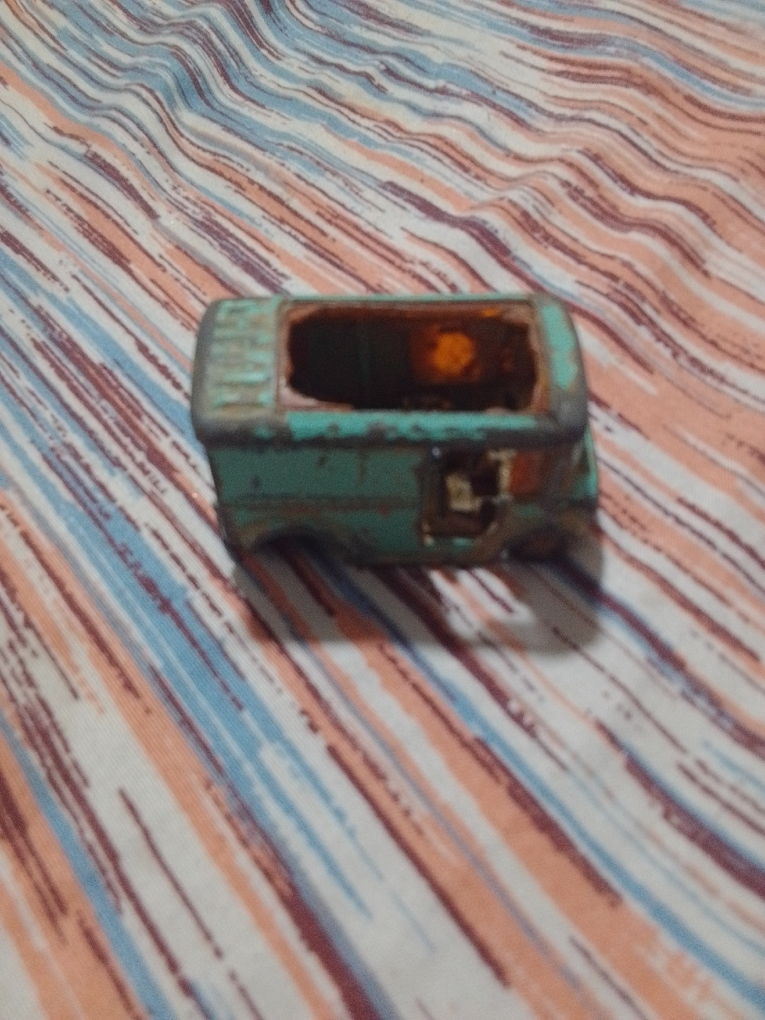 The media file shows a close-up, slightly blurry, overhead shot of a teal-colored toy vehicle, possibly a van, resting on a striped fabric. The fabric features diagonal lines in shades of white, pink, peach, and blue, giving a sense of movement or texture. The toy vehicle is in a state of disrepair, with the top opening clearly visible and appearing to be empty or containing some debris. The paint on the toy is chipped and worn, suggesting it is an older item. The lighting appears to be indoor, with a warm glow emanating from the open interior of the toy, possibly reflecting off something within. No people are visible in the image, and there are no discernible location cues that would definitively place it in La Tagua, Venezuela, other than the provided context. The overall impression is of a neglected toy, likely a childhood relic, set against a visually interesting, textured background.