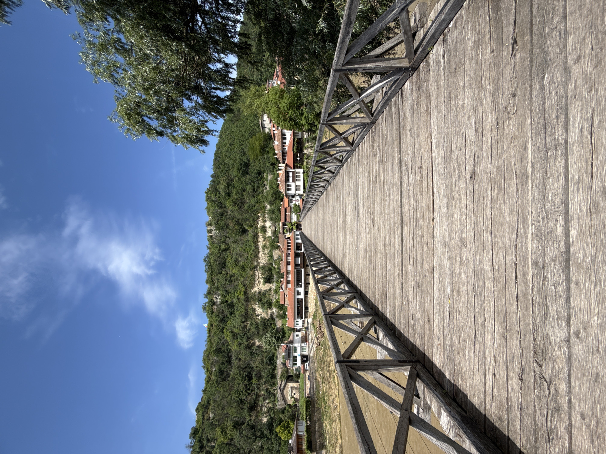 A wooden bridge stretches into the distance in the city of Veliko Tarnovo, Bulgaria. The bridge is constructed of light brown wooden planks and has decorative wooden railings on either side with an X pattern. The bridge leads towards a hillside covered in dense green trees and houses with red tile roofs. The sky is blue with scattered white clouds. A tree with green leaves is visible on the left side of the frame.