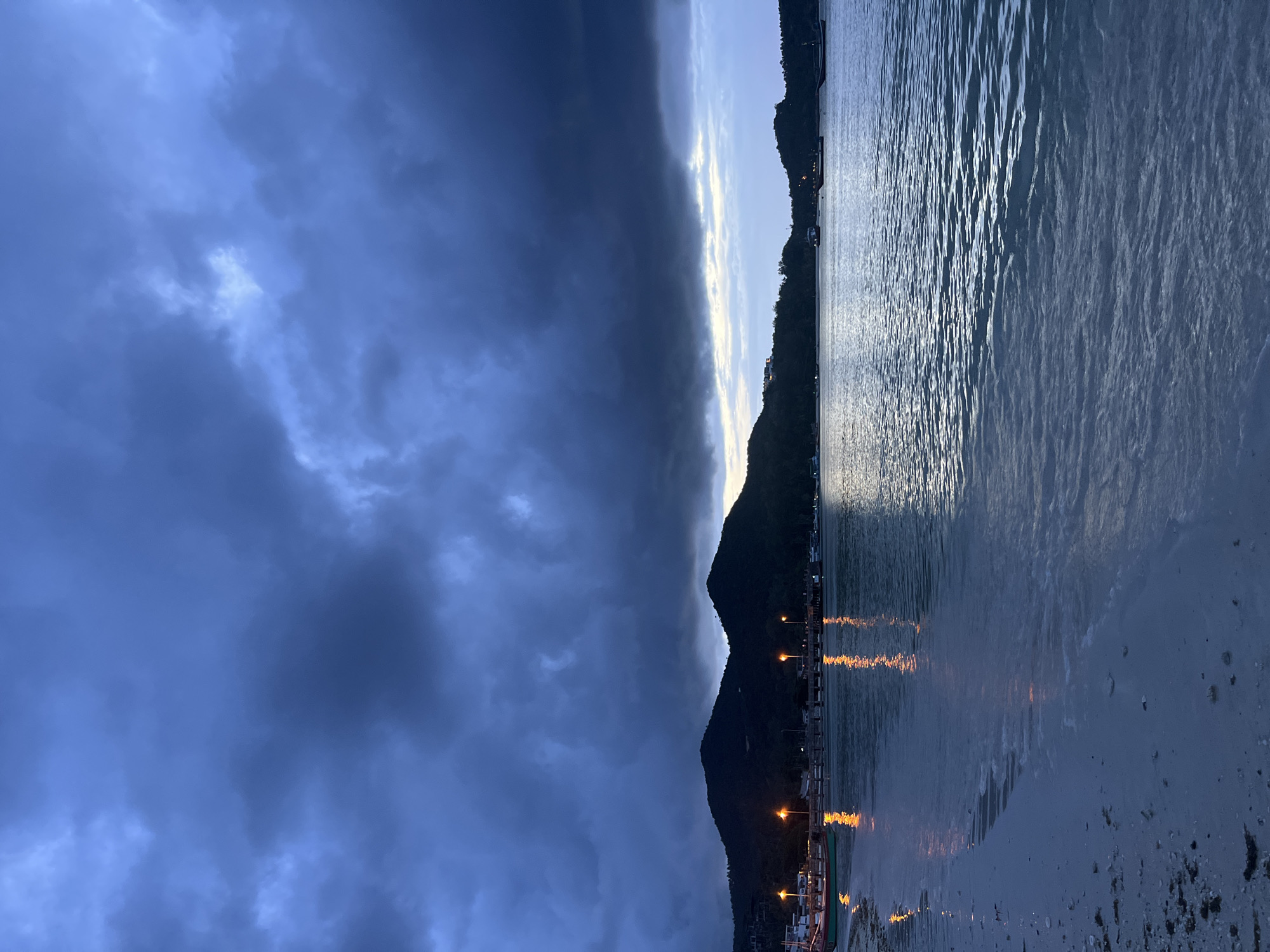 This is an image of a beach and bay in Ko Pha-ngan District, Thailand, at dusk. The sky is filled with dark, ominous clouds, with a sliver of lighter sky visible on the horizon. The water is calm, with gentle waves lapping at the sandy shore. Along the shoreline, there are a few boats, and a pier with several lights casting reflections on the water. In the background, silhouetted mountains rise against the darkening sky. The overall mood is serene and tranquil, with a hint of drama from the cloudy sky.