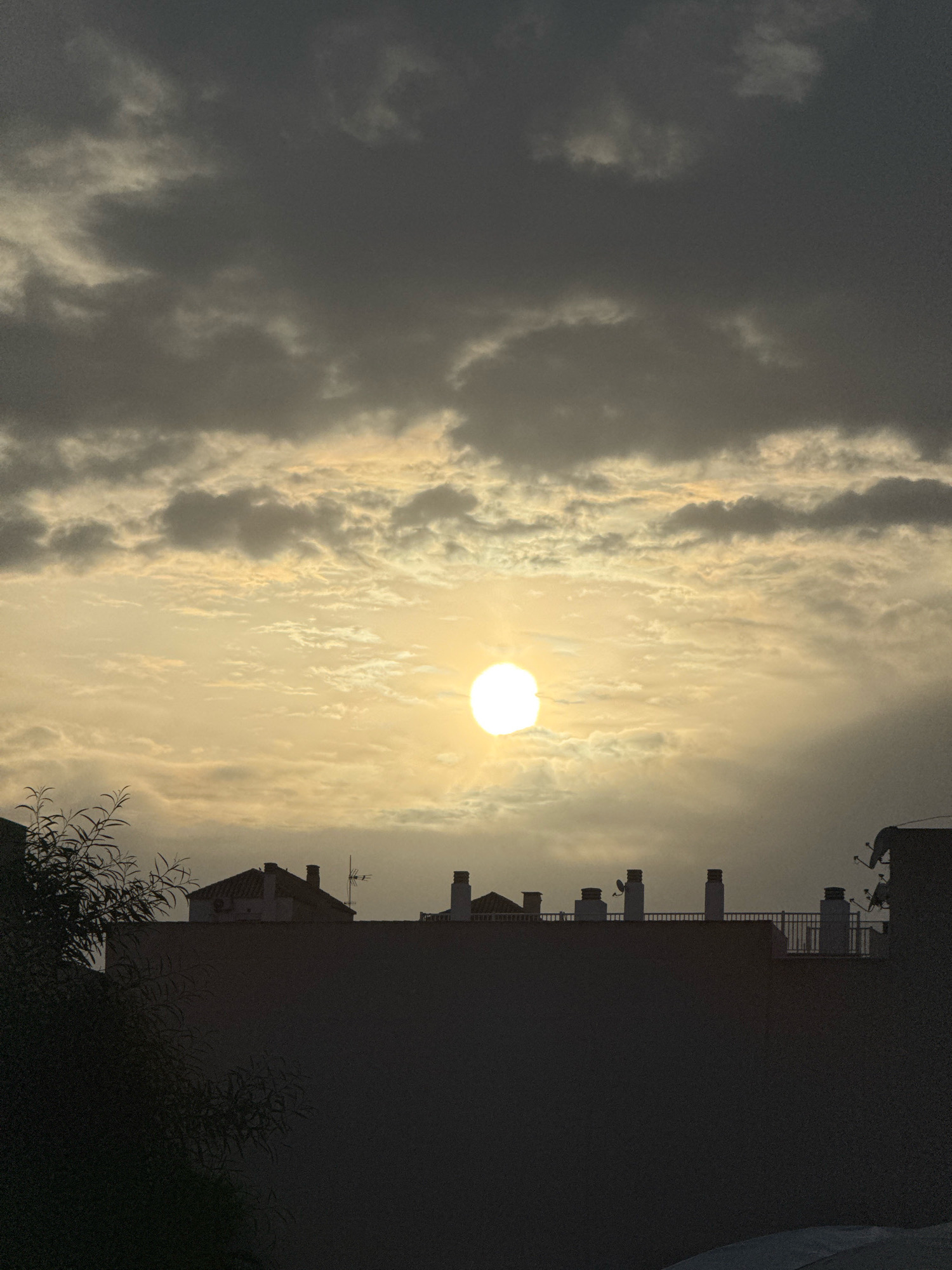 The image depicts a sky with a bright sun obscured by clouds, casting a warm, diffused light over a silhouetted cityscape. The foreground is dominated by the dark shapes of building rooftops and a large, shadowy tree on the left. Numerous chimneys and what appear to be satellite dishes are visible atop the buildings, suggesting an urban environment. The clouds are varied in texture and color, with some areas appearing darker and more ominous, while others are illuminated by the sun, creating a dramatic and somewhat hazy atmosphere. The overall impression is that of a late afternoon or early evening sky over a Spanish city. Given the presence of what are likely satellite dishes, often associated with television reception, and the style of the buildings, it is plausible this scene is located in Torrevieja, Spain. There are no people or active interactions visible in the image.