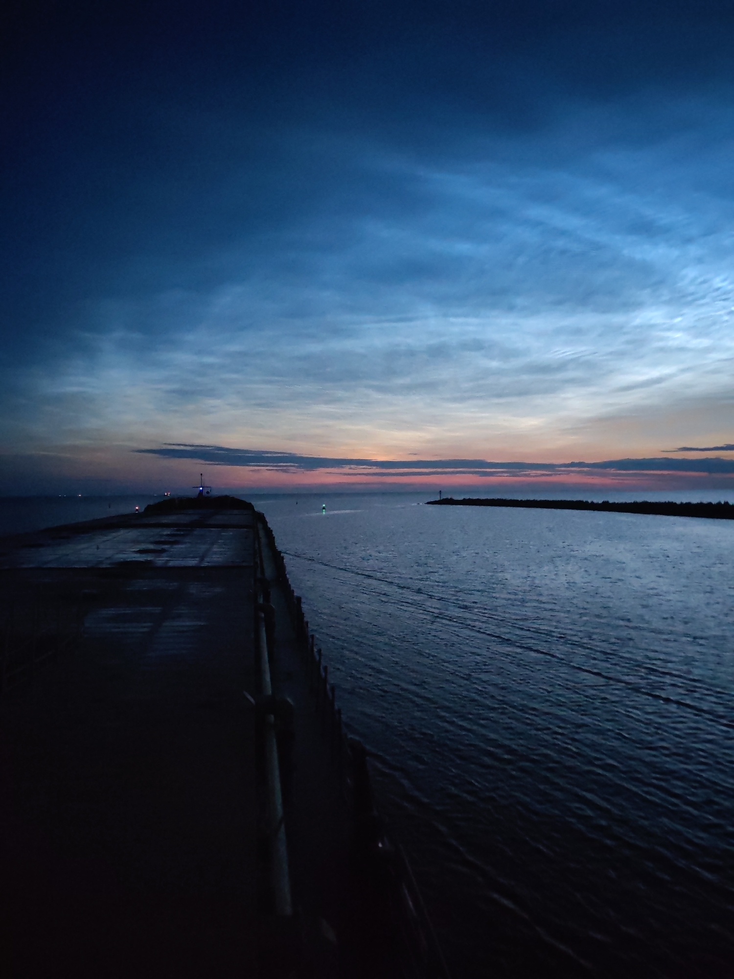 The image depicts a dusky scene at Ozernoe Ust'e, Russia, with a dark, wet pier in the foreground extending into a body of water. The sky above is a deep blue, transitioning to lighter hues towards the horizon where a soft pink and orange glow indicates the lingering twilight. Wispy, luminous clouds, possibly noctilucent clouds, are visible in the upper portion of the sky. The water is calm with subtle ripples, reflecting the dim light. Distant navigational lights, some green and red, mark the waterway, and a faint silhouette of a lighthouse or structure can be seen on the horizon. The overall atmosphere is tranquil and atmospheric, characteristic of the transition from day to night in a coastal or port area. There are no people visible in the image.