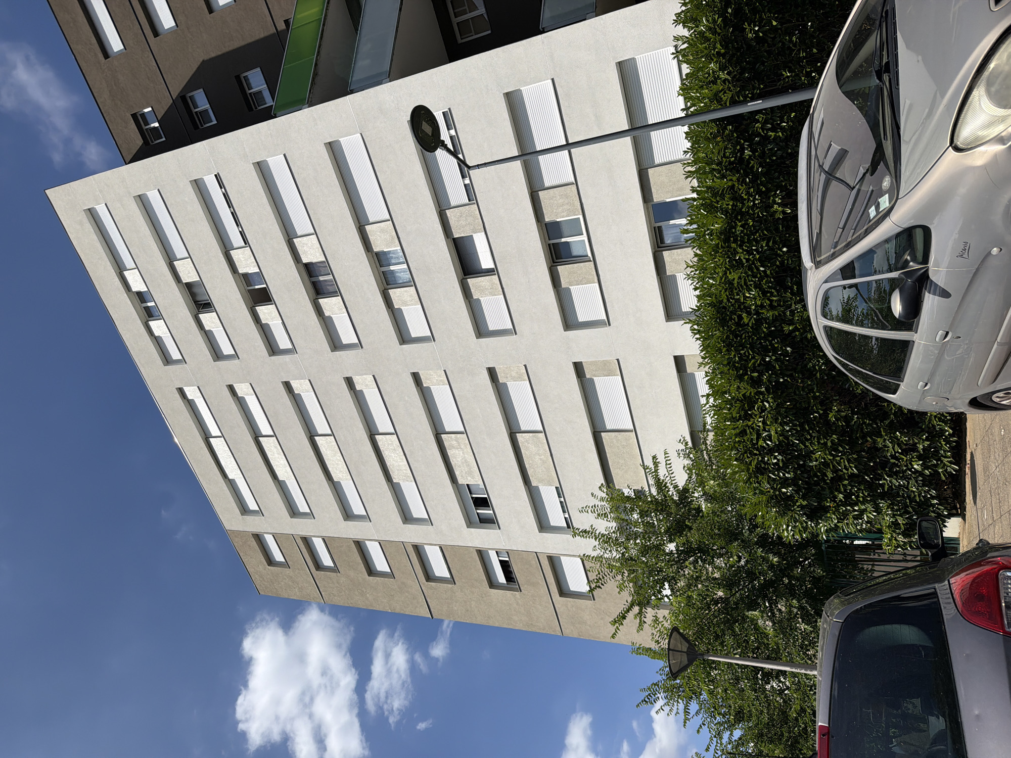 This is a low-angle, full shot of modern apartment buildings in Évry-Courcouronnes, France, under a bright blue sky with scattered clouds. The architecture is characterized by clean lines, alternating light and dark grey facades, and numerous elongated windows with white slatted blinds. The buildings appear to be tall and multi-story.
In the foreground, a silver car is parked on a paved area, partially obscured by a dense green hedge. Further back, a dark grey car is also visible, facing away from the camera. A streetlamp with a triangular head is positioned between the buildings and the cars, extending outwards.
The scene suggests a sunny day, likely late morning or early afternoon, given the strong shadows cast by the buildings. The overall impression is one of urban residential living, with a focus on the architectural design and the interplay of light and shadow. There is no discernible activity or event taking place, and no visible text. The image is captured with a slightly tilted perspective, adding a dynamic feel to the composition.