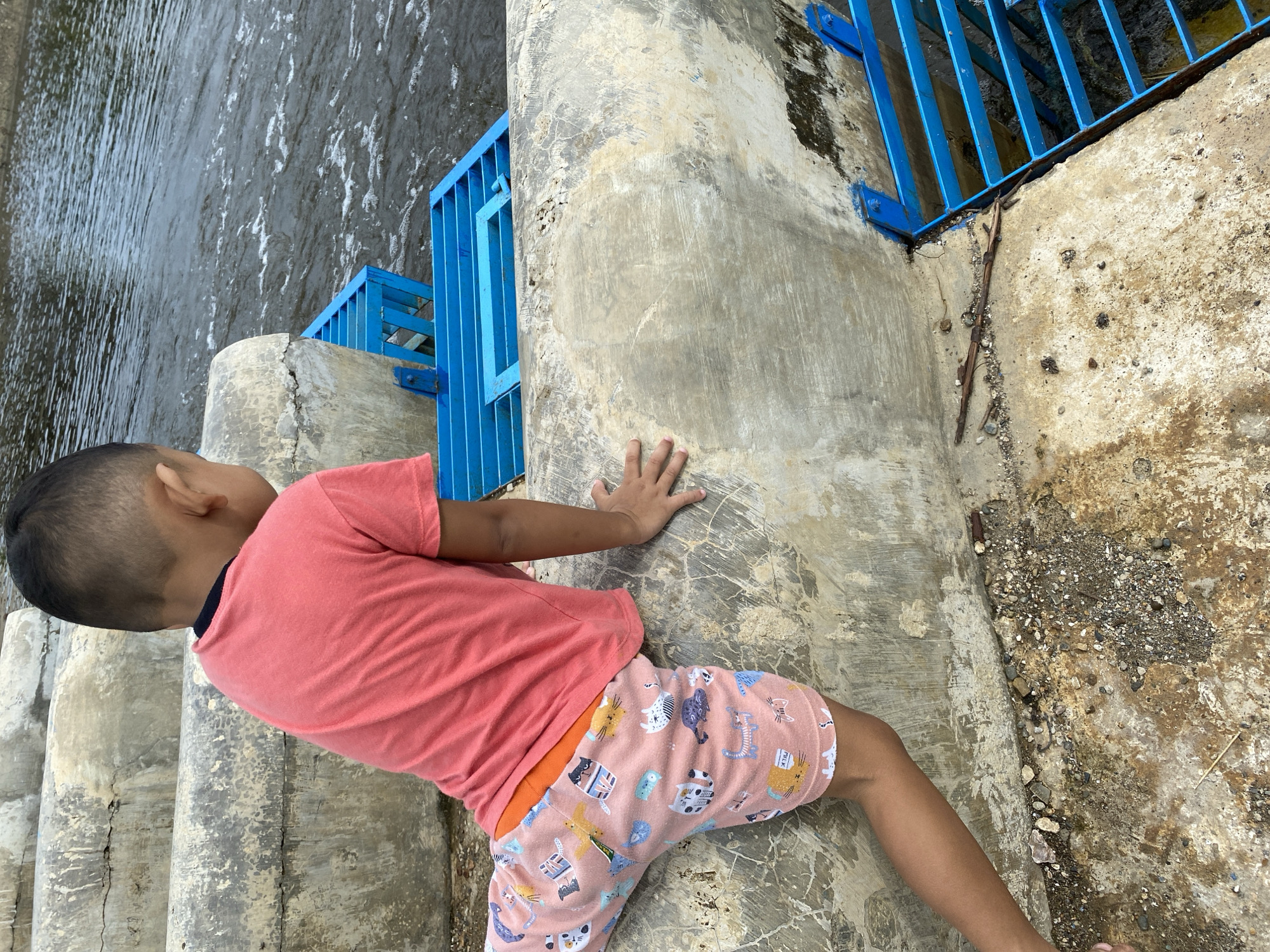 A young boy wearing a coral pink t-shirt and patterned shorts is positioned on a concrete structure near a body of water. The boy is facing away from the camera and towards the water, with his left arm resting on the concrete and his right hand reaching forward. The concrete structure appears to be made of stacked, curved sections, possibly part of a dam or weir. Adjacent to the boy is a bright blue metal grate structure, which sits over the water. The water itself is choppy, indicating some current or movement. The ground beneath the concrete structure is rough, with small stones and dirt visible. The environment suggests an outdoor, possibly recreational or functional area, in Aneuk Paya, Indonesia.