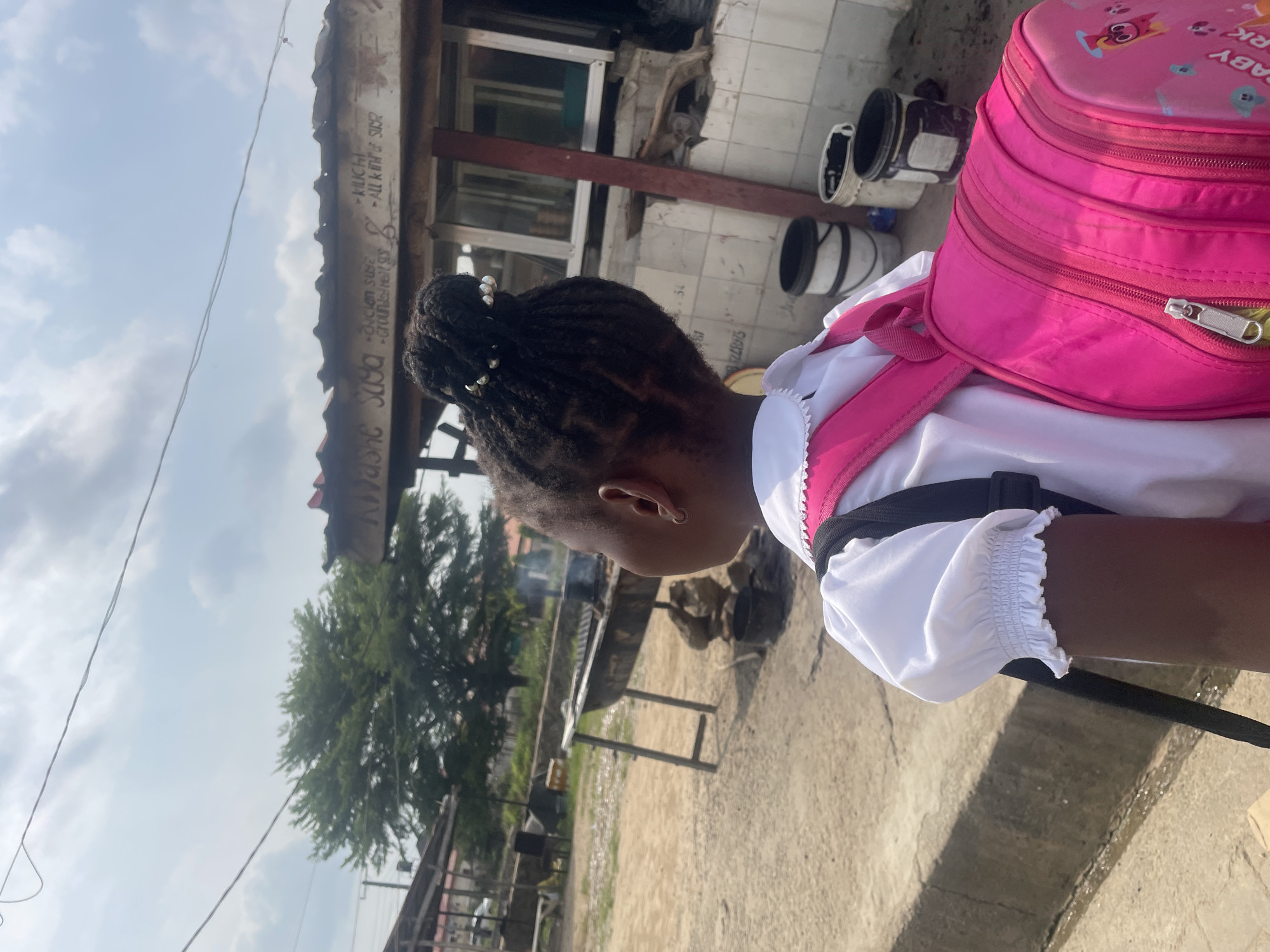 The image features a young girl from behind, facing to the left, with a bright pink backpack on her shoulders. She is wearing a white blouse with ruffled sleeves. The backpack is adorned with cartoon characters. The setting appears to be an outdoor area with a dusty, concrete ground and some vegetation in the background. There's a building with a corrugated metal roof in the middle ground. Some white and black buckets are visible near the building. The sky above is partly cloudy, suggesting it's daytime.
There is visible text on the metal roof, though it is partially obscured and difficult to read clearly. Some discernible words include "CHICKEN SUSE," "-GROUND HEIL SUGE," and "MUCHET - ALL KINDA SUGE." The overall impression is of a casual, possibly residential or commercial outdoor space in Abuja, Nigeria, with the child possibly on her way to or from school, given the backpack. The lighting indicates it's likely late morning or early afternoon.