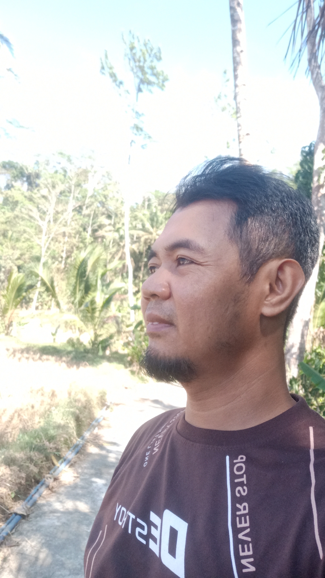 In an outdoor setting, a man of Asian descent is captured in a profile view, looking to the left. He has short, dark hair, a trimmed beard, and his skin appears to be tanned. He is wearing a dark brown t-shirt with white lettering that appears to be in a stylized font. The background is blurred, but it suggests a tropical environment with lush green foliage and palm trees. The sky above is bright blue with a few white clouds, indicating a sunny day. The ground in the foreground is a concrete path, and to the left of the path, there is dry grass and some kind of blue pipe or conduit. The image is taken in Desa Burujul Jaya, Indonesia.