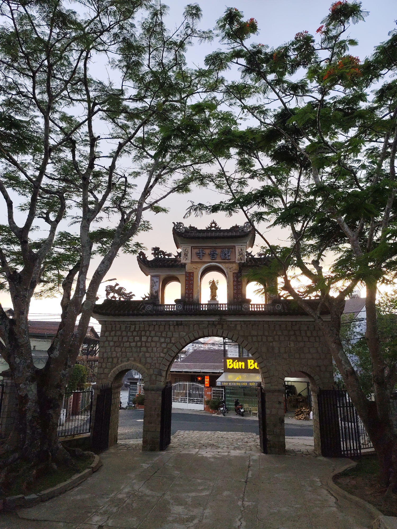 This image captures a serene view of a traditional Vietnamese archway and an ornate structure, framed by two large, verdant trees, set against a warm-toned sky. The scene is likely in Da Lat, Vietnam, as indicated by the context.
The main subject is a substantial stone archway, constructed from rough-hewn blocks, featuring two arched passages. Above this stone base, an elaborate, multi-tiered structure with a traditional Asian roof stands. This upper structure boasts multiple arched openings; a prominent statue, possibly a deity or cultural figure, is visible within the central arch, dramatically back-lit by the sun. The sun's warm glow, suggesting either sunrise or sunset, illuminates the figure and the sky, creating a soft, ethereal outline around the entire upper edifice.
Large trees, possibly Flamboyant trees with hints of reddish-orange blossoms, dominate the left and right sides of the frame, their intricate branches and dense green foliage arching over the structure. Through the open passages of the stone archway, a glimpse of a street in Da Lat is revealed. Visible details include buildings, two parked motorcycles, and a glowing yellow business sign displaying the Vietnamese text "Bún Bò," with smaller, less clear text below that appears to be "BÁNH MÌ XÍU MẠI CHÉN" and "NƯỚC...". Metal gates are partially visible within the archway openings.
The time of day is either early morning or late afternoon, characterized by the soft, warm light and the strong backlighting effect on the statue. The sky is clear, indicating fair weather. No people or specific events are depicted; the image captures a tranquil moment at what appears to be a significant cultural or religious landmark.
