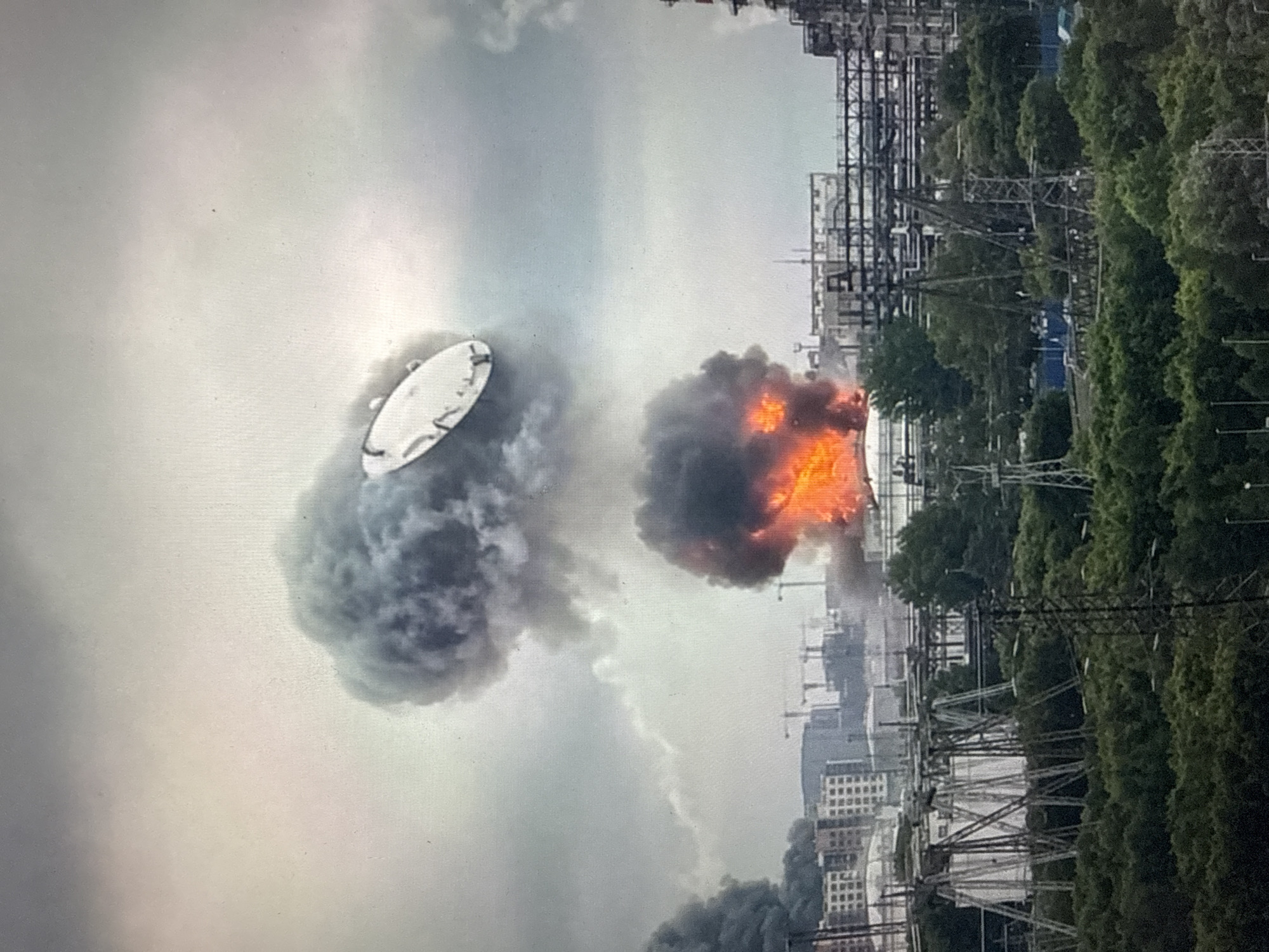 In the city of Unknown, Unknown, a fire erupts at an industrial facility, sending thick plumes of dark smoke into the overcast sky. The flames are visible in the lower center of the frame, consuming a portion of the structure. A large, disc-shaped object is suspended in the air above one of the smoke clouds. The industrial complex features numerous pipes, storage tanks, and scaffolding. In the background, several buildings and construction cranes suggest a developing urban or industrial area. A dense line of trees forms the foreground, obscuring the lower parts of the facility. Electrical towers are also visible within the industrial landscape. The scene is chaotic, with the fire and smoke dominating the visual.