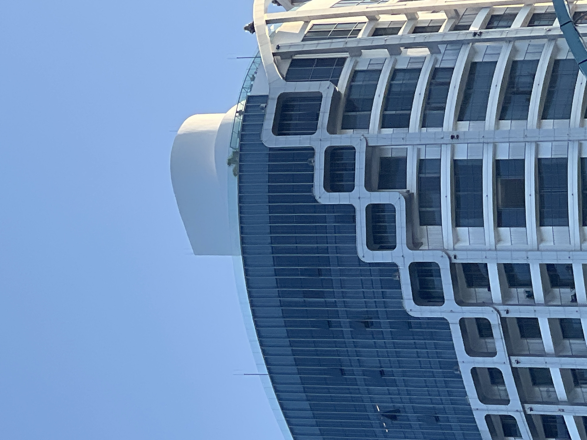 The image displays an upward view of the APEC Center, a modern high-rise building located on Pham Van Dong Street in Nha Trang, Khanh Hoa, Vietnam, under a clear blue sky.
The building's architecture is characterized by two distinct vertical sections. The left section features a prominent, curved dark blue glass curtain wall with vertical divisions, showcasing several partially open windows. Above this glass facade, a rooftop area with a transparent glass railing contains sparse green vegetation and two indistinct human figures. This section culminates in a large, smooth, white, rounded dome-like structure at the building's apex.
The right section of the building presents a white facade with multiple horizontal levels of curved balconies, some equipped with railings. This white section is visibly integrated with the glass facade by a lighter-colored, grid-like architectural element featuring rounded square openings. On a roofline at the far upper right, two small, dark, unidentifiable objects or figures are present. Multiple thin, pole-like structures, consistent with antennae, project from various rooftop elevations. The scene is illuminated by natural light, indicating daytime conditions.