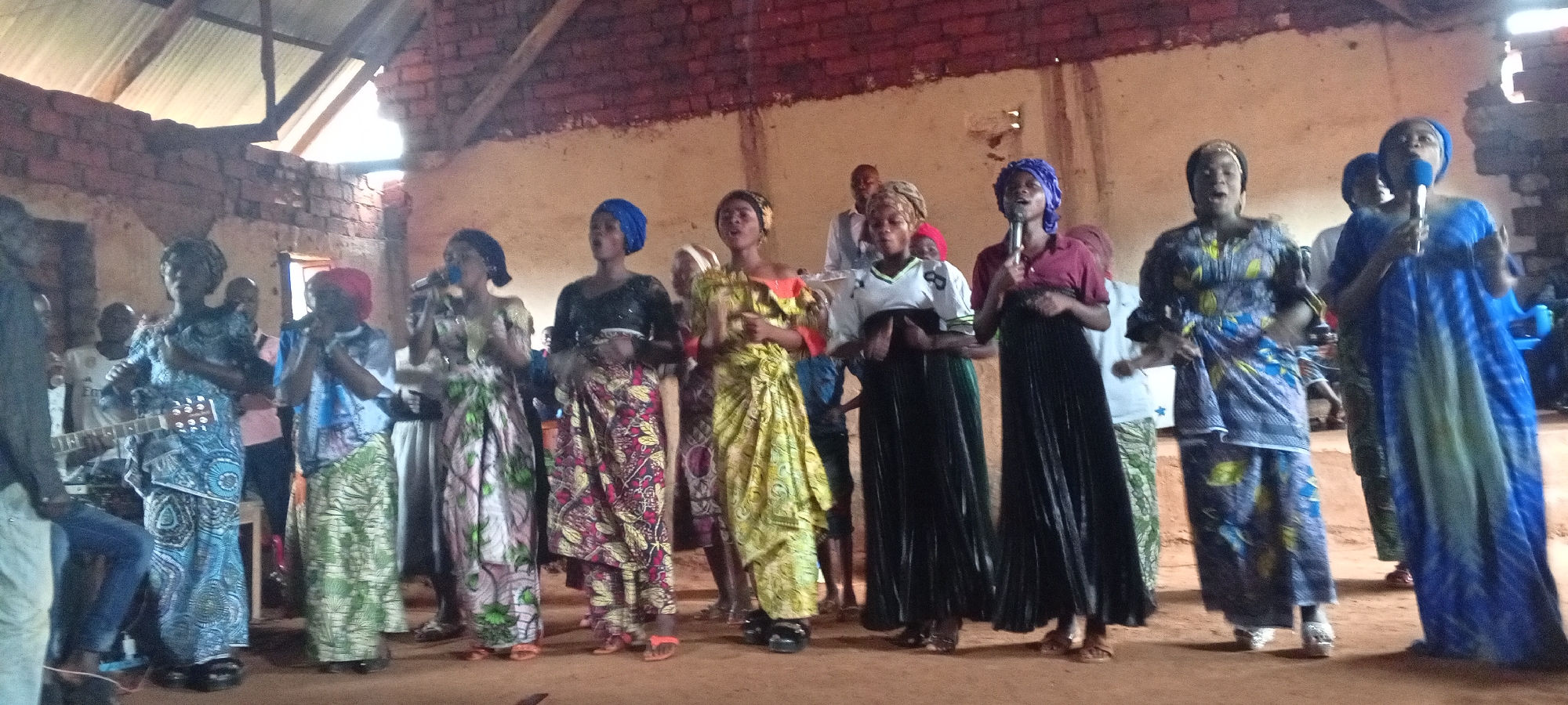 A group of approximately ten people stand in what appears to be a church or community center in Ngalula, Democratic Republic of the Congo. The group consists primarily of women, several of whom are holding microphones and appear to be singing or speaking. They are dressed in a variety of colorful patterned fabrics, some wearing head coverings. A man to the left plays a guitar. The interior is sparsely decorated with exposed brick or mud walls. The floor is dirt or packed earth. Light enters through openings in the roof. The overall impression is of a gathering or performance in a simple, perhaps rural, setting.