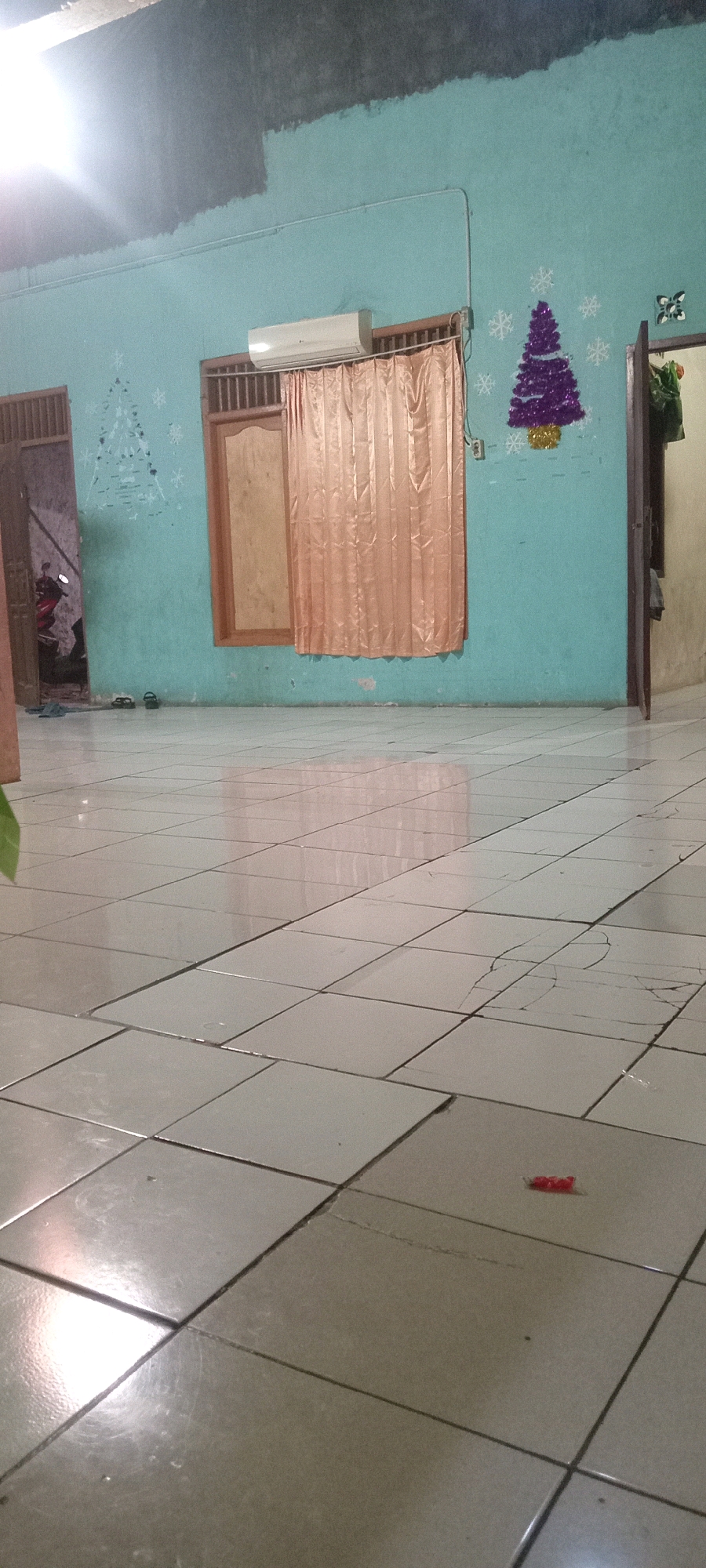 The image depicts an interior scene, likely a residential dwelling, in Depok, Indonesia. The room features turquoise-painted walls with a dark, unfinished section at the top. Decorative elements on the wall include a Christmas tree with purple tinsel and white snowflake decals, as well as a Christmas tree outline made of white decals. An air conditioning unit is mounted above a window covered by peach-colored curtains. A wooden door or window frame is visible to the left, leading to an area where a motorcycle is parked. The floor is tiled with light-colored square tiles, some of which show cracks and wear. A small red object, possibly a piece of candy or a toy, lies on the floor in the foreground. The lighting suggests it may be daytime, with sunlight reflecting off the tiled floor. The overall impression is of a simple, lived-in space, possibly decorated for a holiday.