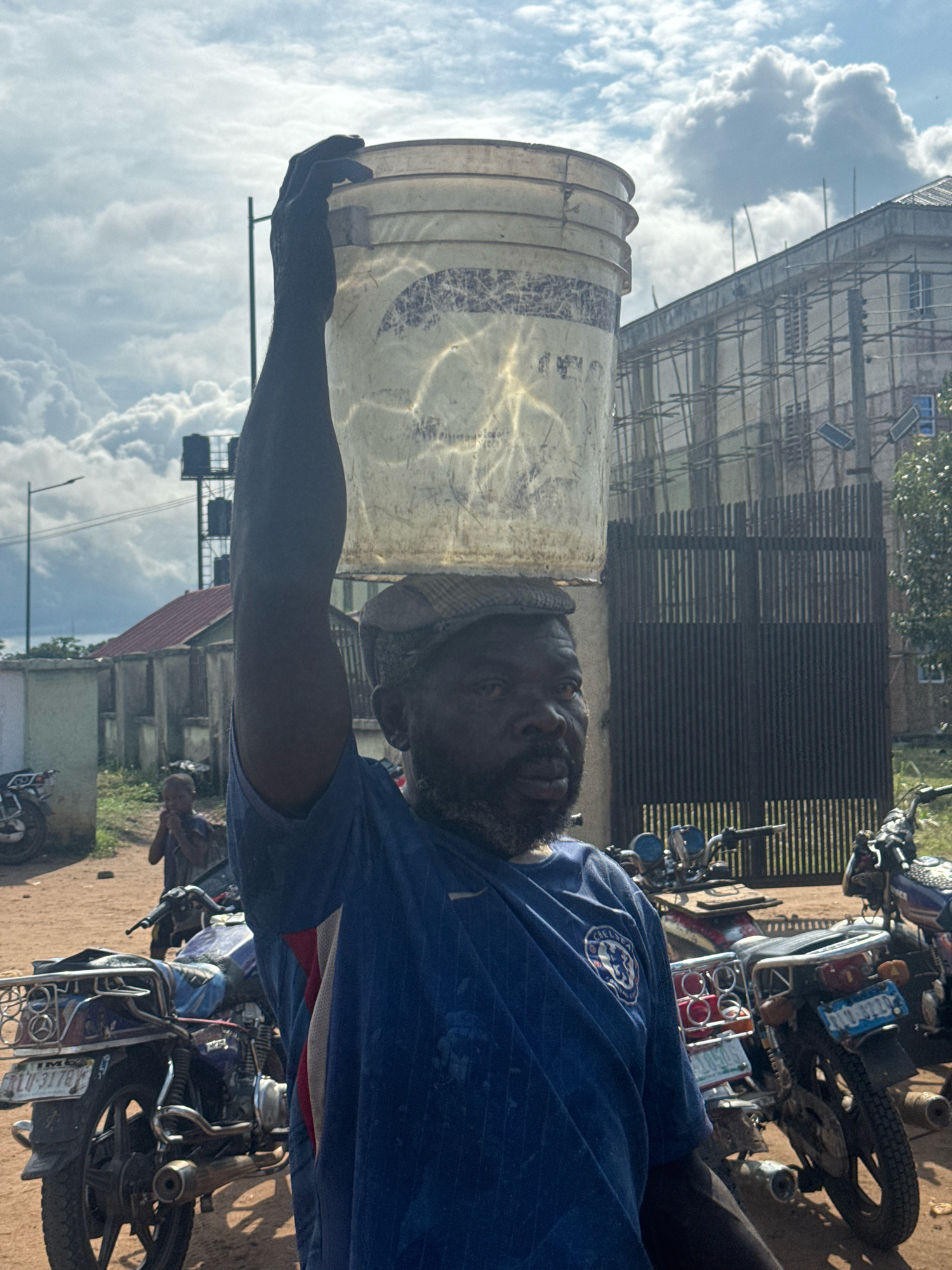 A man is carrying a bucket on his head, wearing a blue t-shirt with a Chelsea logo on the left chest. He is wearing a cap and has a beard. He is standing in what appears to be a dusty street in Ihiagwa, Nigeria. In the background, there are several motorcycles, a partially constructed building with scaffolding, a tall structure with water tanks, a fence, and some houses. A child is visible in the background, standing to the left of the man. The sky is partly cloudy.
