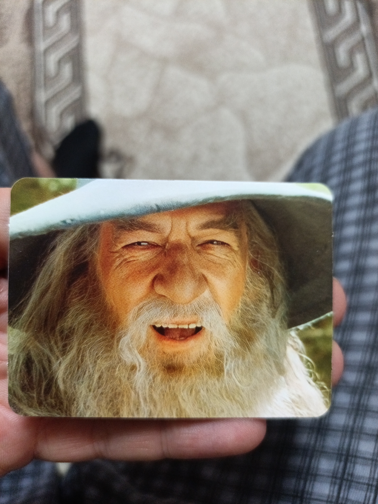 The image shows a close-up of a person's hand holding a card that features a portrait of Gandalf the Grey, a character from The Lord of the Rings. The card is rectangular with rounded corners and depicts Gandalf's face, complete with his signature hat, long white beard, and a wide smile. The background of the image shows a blurred pattern of fabric, likely clothing or upholstery. There is no indication of the city of Nyagan, Russia, in this image.