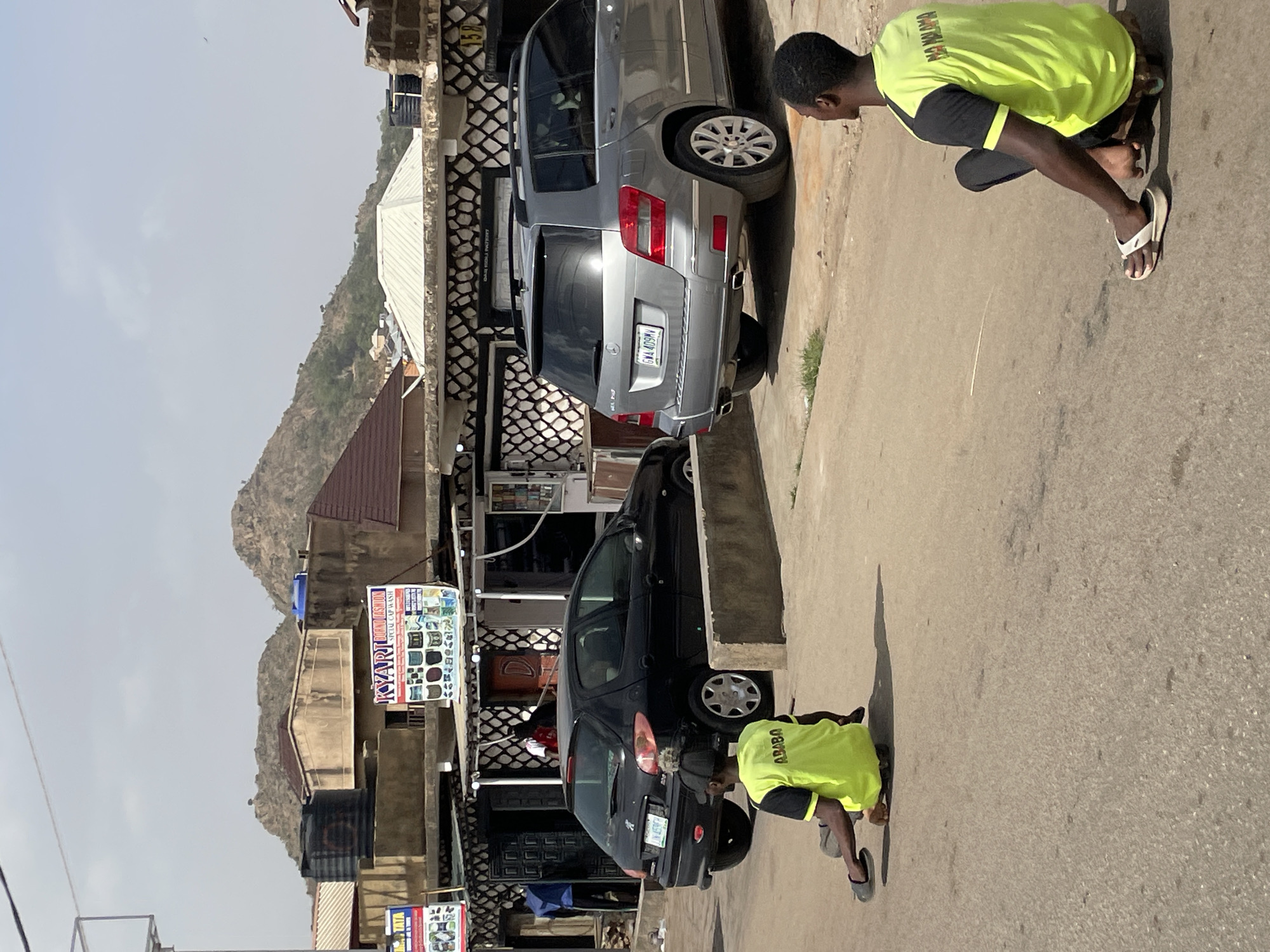 The image captures a street scene in what appears to be a town or city, consistent with Jos, Nigeria, featuring two men, two cars, and buildings against a backdrop of rocky hills.
Two men, both dressed in matching neon yellow/green t-shirts with black sleeves, are crouching on a paved road. One man is in the foreground, positioned near the rear of a black Peugeot hatchback. The second man is further back, near the rear of a silver Mercedes-Benz GLK-Class SUV. Both appear to be inspecting or working on something low to the ground, possibly related to the vehicles or the roadside.
The clear, bright sunlight casts distinct shadows, suggesting it is midday or early afternoon with a clear sky. The buildings lining the street exhibit local architecture, and a prominent sign on one building reads "K'YARI MICRO..." with "SPECIA, CAP & WASH" below, displaying images of electronics or household items. Another partially visible sign mentions "SUPERMARKET." The number "150" is also visible on a patterned building wall.
Visible text on the men's t-shirts includes "MAWUOMA" on the back of the man by the Mercedes and "BABO" on the back of the man by the Peugeot. The silver Mercedes-Benz has a Nigerian license plate "AA 609 HY," while the black Peugeot's plate is partially visible as "KUJ-771...". The background features characteristic rocky, arid hills, reinforcing the Nigerian context.