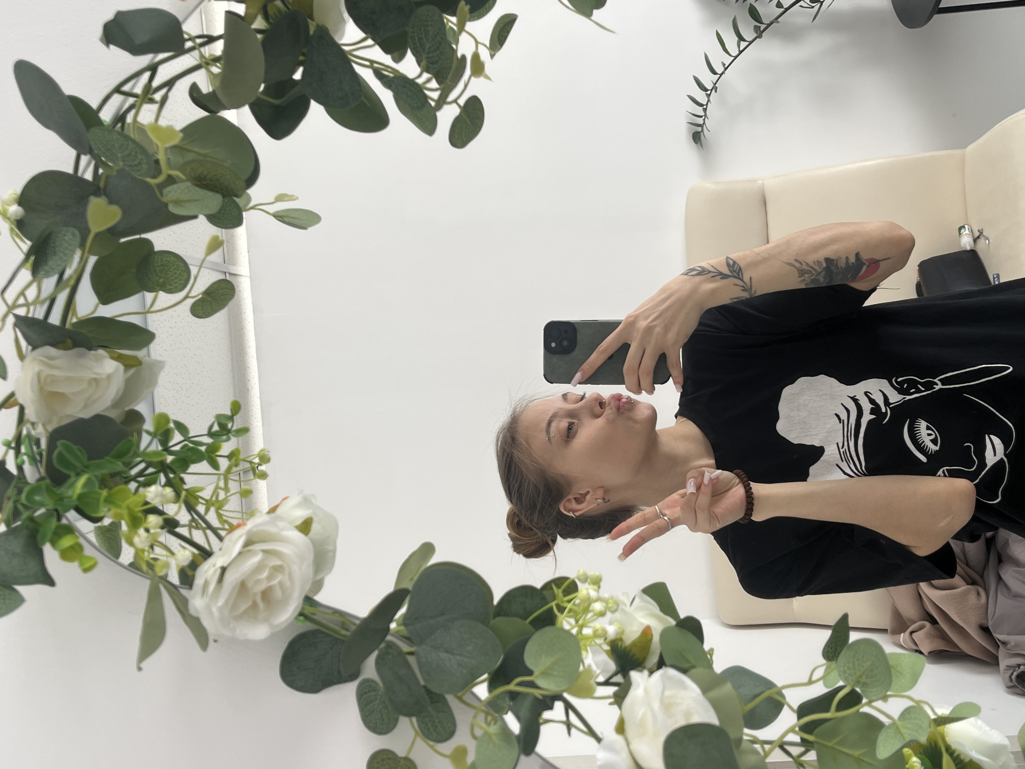 A mirrored selfie captures a young woman in what appears to be an indoor setting. The woman holds a smartphone in her right hand, taking the picture in front of a mirror adorned with floral decorations consisting of white roses and greenery. She is making a peace sign with her left hand and posing with pursed lips towards the phone. She is wearing a black t-shirt with a white graphic design, and her hair is styled in a bun. Tattoos are visible on her left arm. A beige couch is partially visible behind her. The location appears to be in a brightly lit room with white walls and a clean aesthetic. The absence of recognizable architectural features prevents definitively establishing this location as Фоки, Russia.