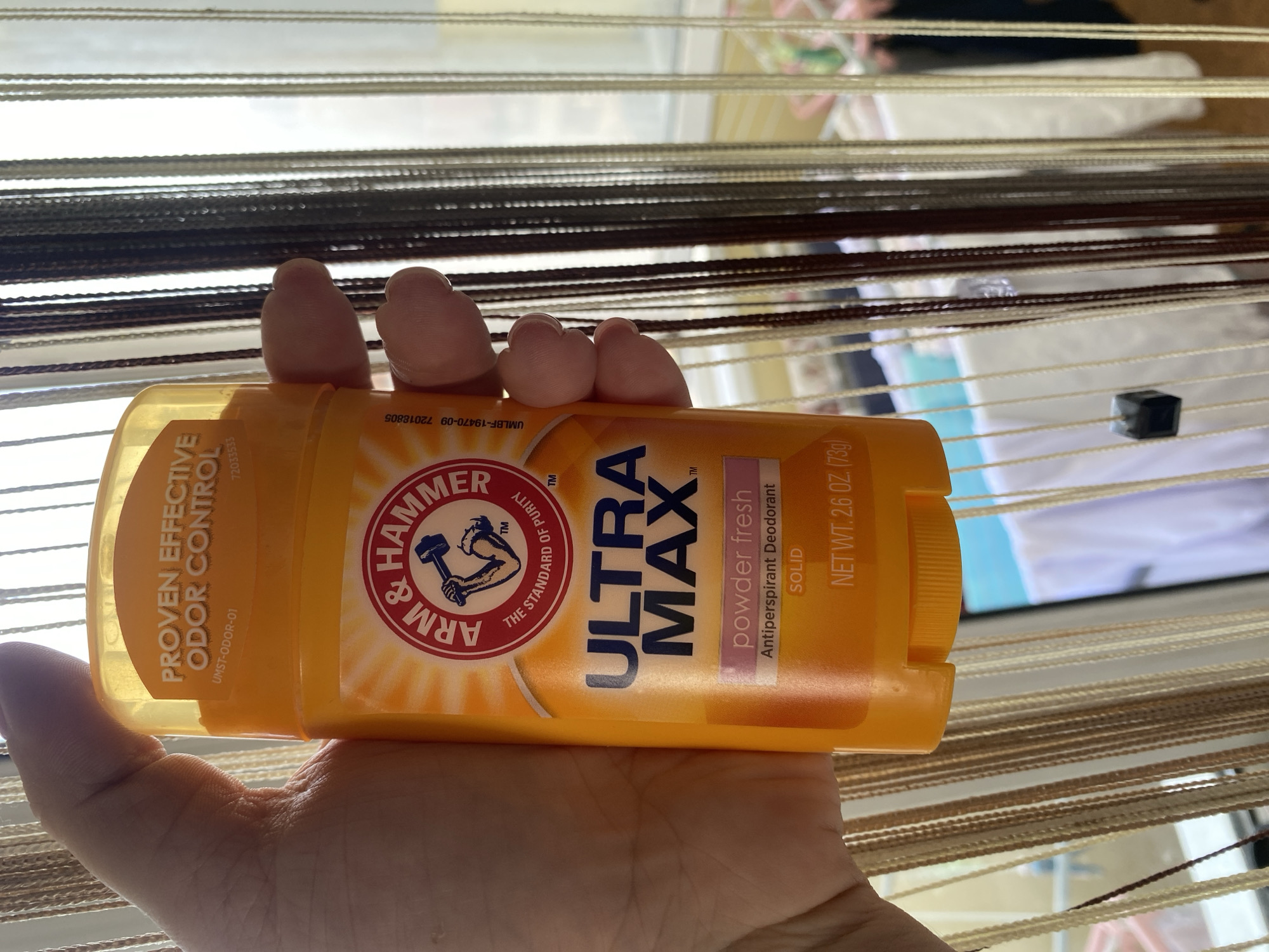 A hand holds an orange deodorant stick, the Arm & Hammer Ultra Max brand, in front of a window with vertically hanging, multicolored curtains. The deodorant has a yellow cap and features the Arm & Hammer logo, which depicts a muscular arm holding a hammer. The text on the stick indicates "PROVEN EFFECTIVE ODOR CONTROL," "ULTRA MAX," "powder fresh," "Antiperspirant Deodorant," "SOLID," and "NET WT. 2.6 OZ. (73g)". The background is blurred but suggests an indoor setting with sunlight filtering through the window and curtains. There are no people or significant objects in the city of Chornomorsk, Ukraine, that are visible in this image.