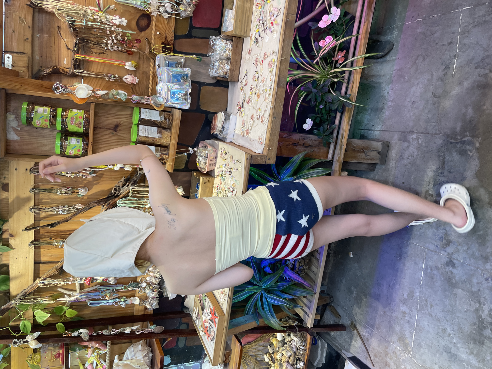 In Phan Thiết, Vietnam, a person wearing a white hooded top and American flag-themed shorts stands in front of a display of shell jewelry and other souvenirs. The person is facing away from the camera, with their left arm raised as if examining a necklace. The background features wooden shelves laden with various trinkets, including jars of nuts or dried goods, packaged jewelry, and a collection of shells. The display is set up outdoors, with plants visible in the foreground and a concrete floor. The lighting appears to be a mix of natural and artificial, with colorful lights casting a purplish and green hue on the floor.
