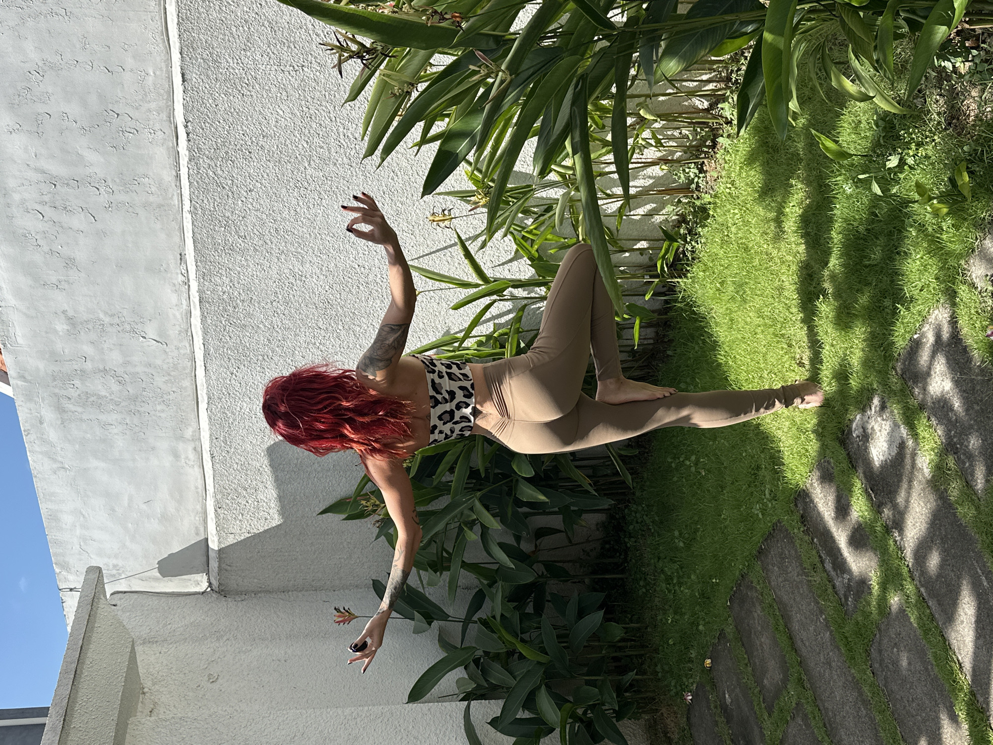 A woman with bright red hair is standing in a yoga pose called Vrikshasana, or Tree Pose, in a sunny outdoor courtyard. She is barefoot and wearing a leopard print tube top and beige leggings. Her right leg is bent, with her right foot resting on her inner left thigh, and her left leg is extended straight down. Her arms are outstretched to the sides, with her palms facing up and her fingers in a mudra. The courtyard has a white textured wall in the background, with lush green foliage and plants growing along the wall. There are also stepping stones and grass in the foreground. The scene is lit by bright sunlight, casting shadows on the ground. The environment appears to be a private residential area, possibly a villa or a home, in the city of North Kuta, Indonesia.