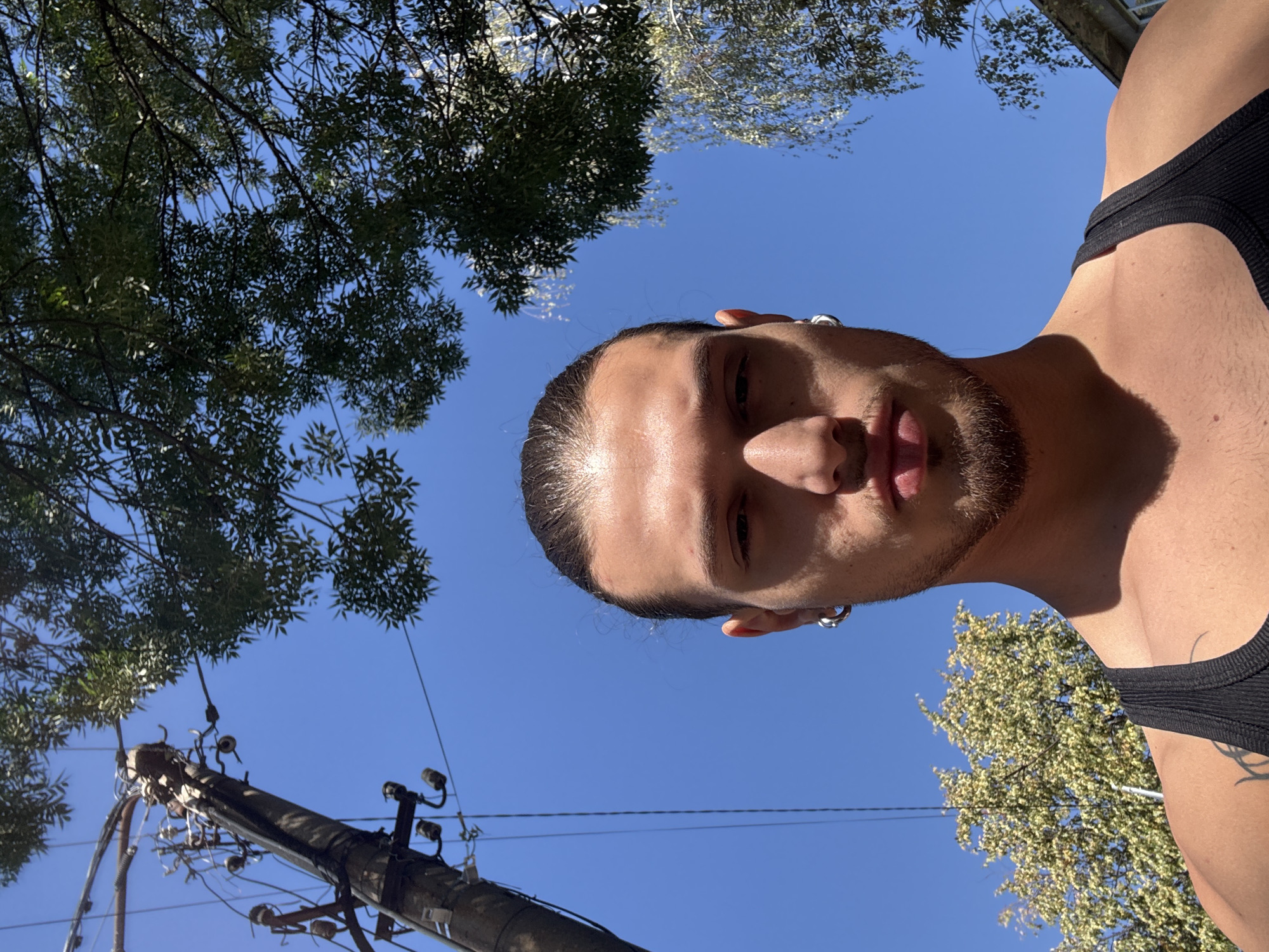 The image shows a low-angle, close-up shot of a young man with dark hair pulled back, wearing a black tank top and silver hoop earrings. He has a light beard and is looking towards the camera. Behind him, the sky is a clear blue, with the top of a utility pole and leafy green trees visible. The scene is likely outdoors. The image does not provide enough information to determine the location in the city of Sofia, Bulgaria.