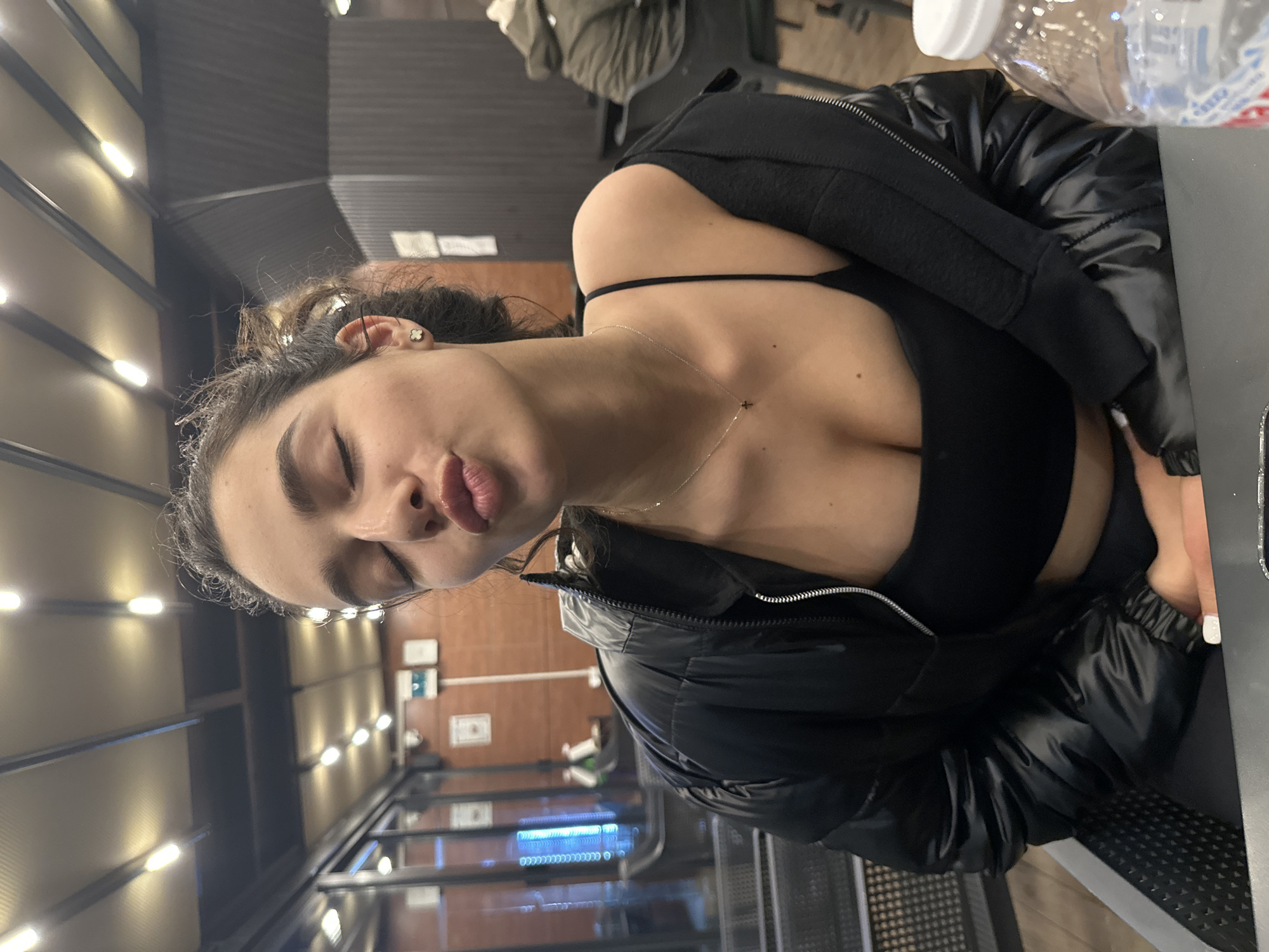 A woman with dark hair and fair skin is depicted from the chest up, indoors. She wears a black bra, a black puffer jacket, and a delicate gold necklace. Her eyes are closed, and her lips are pursed. She is seated at a table next to a clear plastic water bottle. In the background, there is a well-lit interior with brown wood and metal structures resembling gym equipment. The location is possibly a gym or fitness facility in Plovdiv, Bulgaria, given the industrial-style lighting and equipment visible.
