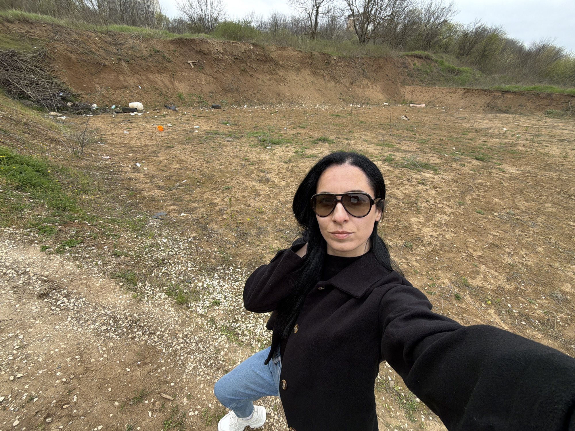 The image shows a woman with dark hair and sunglasses in an outdoor setting. She is wearing a black coat, blue jeans, and white sneakers. The background appears to be a dirt area with some vegetation and an earthen berm. Scattered trash is visible in the background. The location appears to be an informal shooting range or dumping ground. It is difficult to definitively pinpoint the location as Pleven, Bulgaria without additional context.