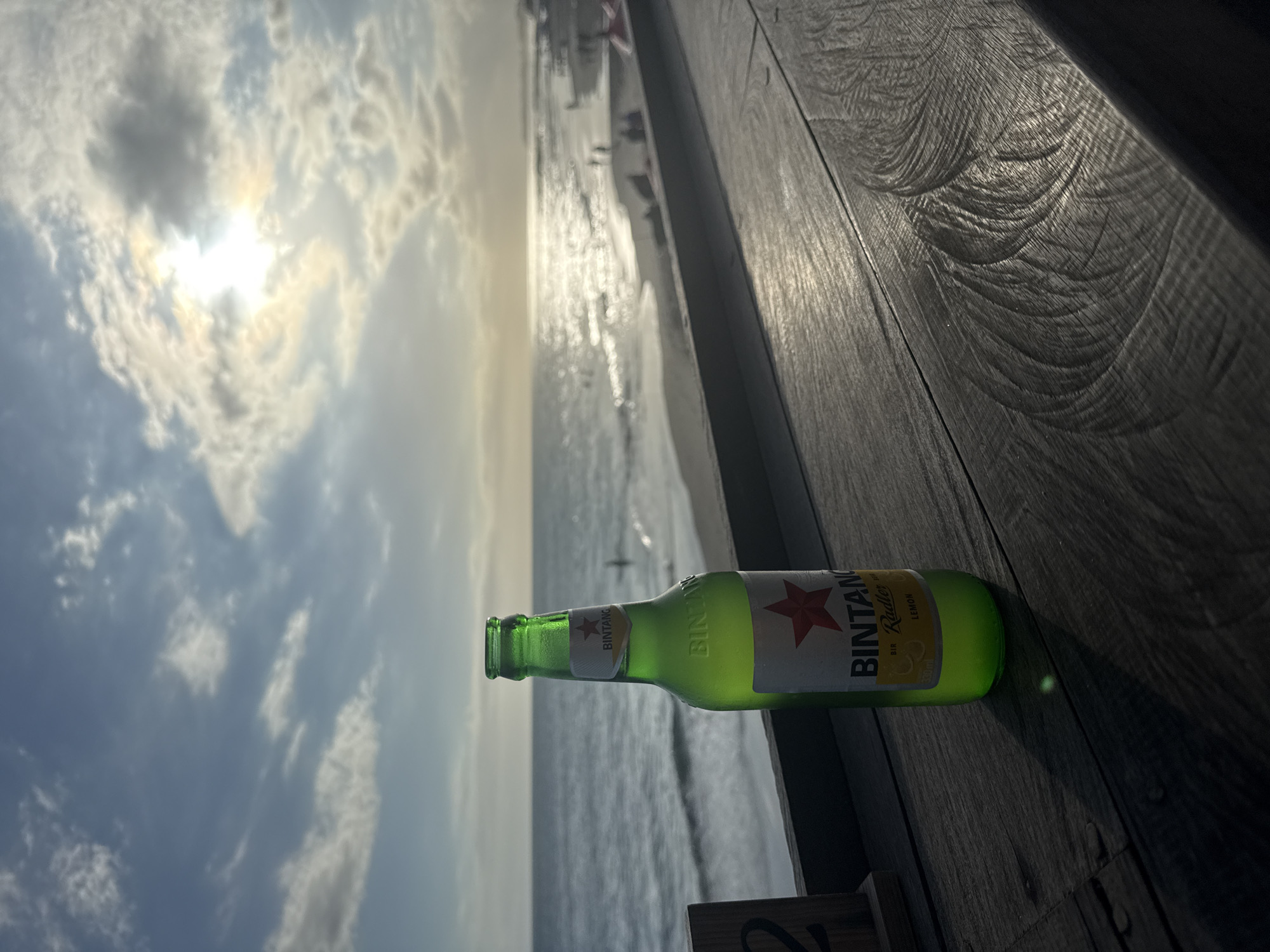 The image depicts a Bintang Radler beer bottle lying on a wooden surface. In the background, there's a beach scene with the sea, sky, and some clouds. The sun is visible through the clouds. The location is identified as North Kuta, Indonesia. There are some people in the beach.