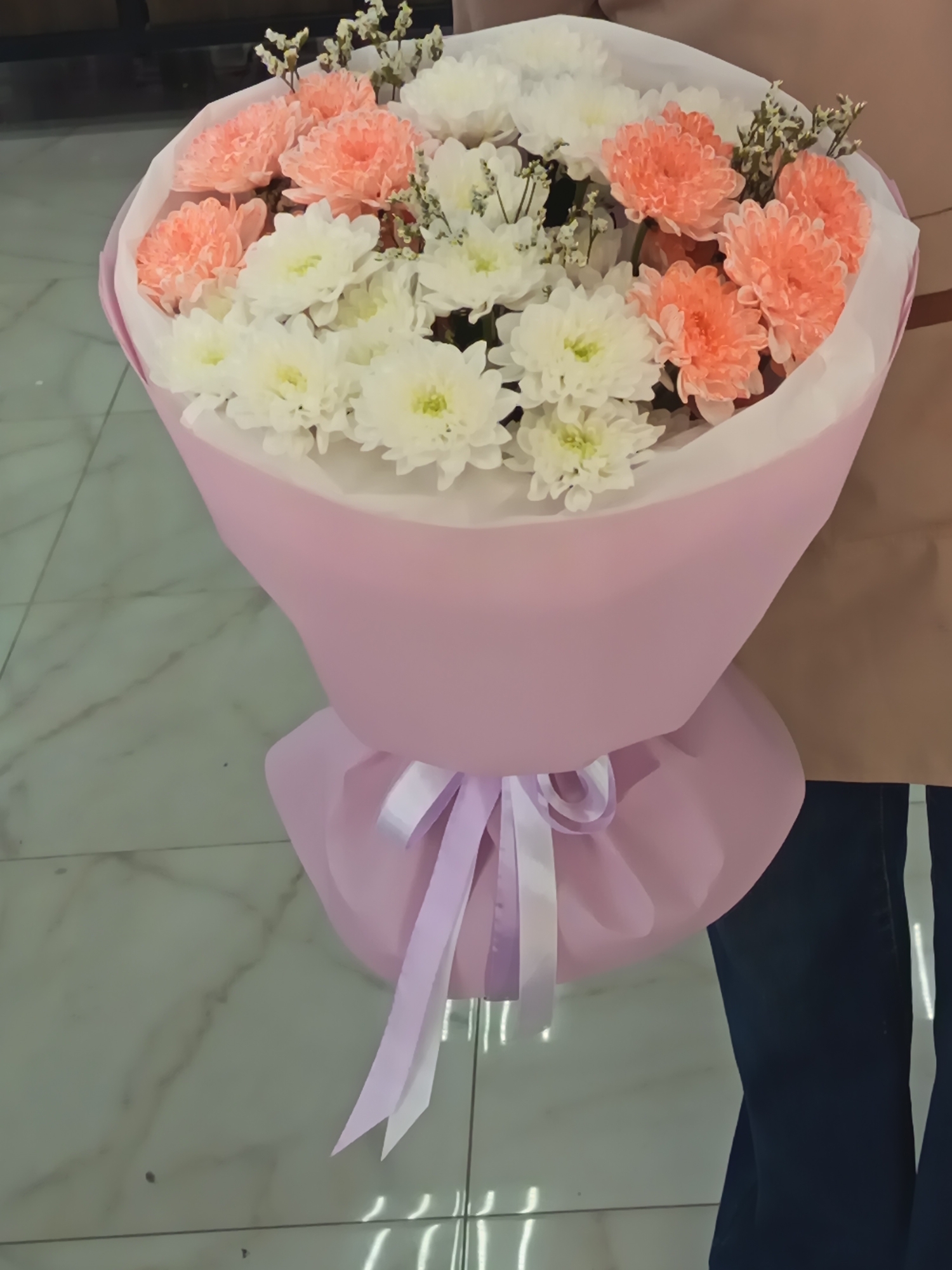The image shows a bouquet of flowers, with white and peach-colored chrysanthemums. The bouquet is wrapped in pink paper and tied with a light purple ribbon. The background includes a tiled floor and part of a person wearing jeans. It is unlikely to determine that the picture was taken in the city of Nizhnevartovsk, Russia, based on the visual elements present.