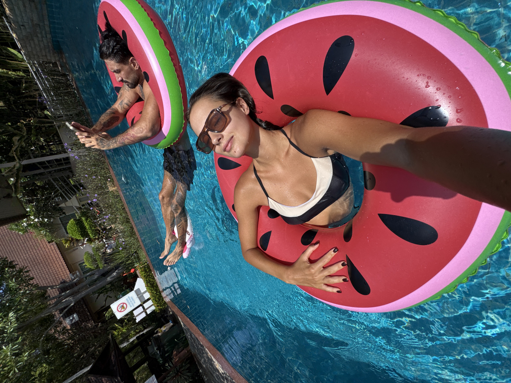 Two individuals, a male and a female, are in a swimming pool using watermelon-themed inflatable rings. The female wears a black and white swimsuit and sunglasses, and her hand rests on the red inflatable. The male, tattooed, is partially submerged and holding a mobile phone. The pool is located at a resort or residence in Pattaya City, Thailand, as indicated by the signage and building visible in the background. The scene includes foliage and part of a building with a red tile roof.