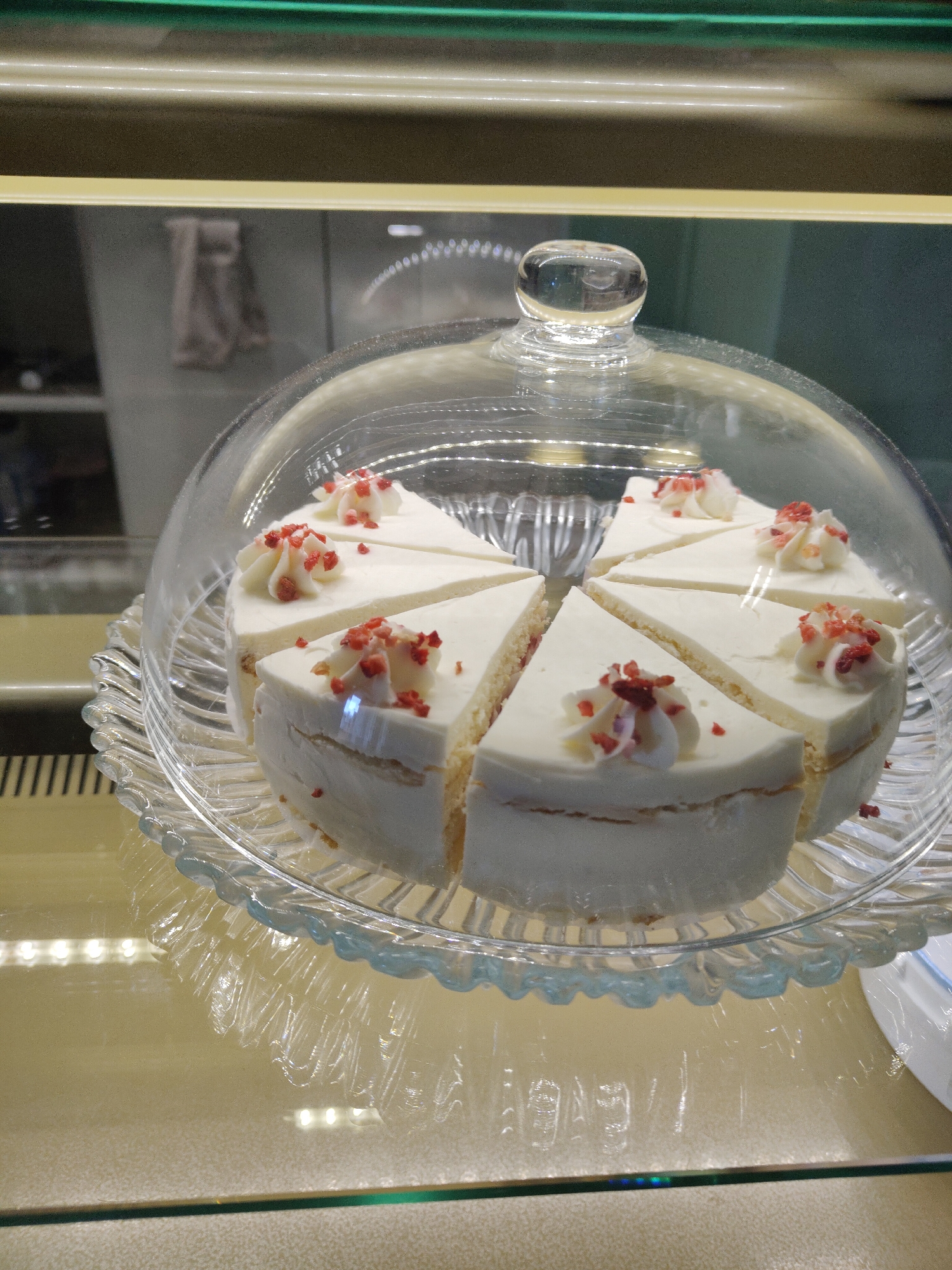 A sliced cake, topped with white frosting and red sprinkles, is displayed on a clear glass pedestal dish with a domed cover in a refrigerated display case. The location is Unknown, Unknown. The cake is cut into eight slices. The display case is illuminated by fluorescent lighting.