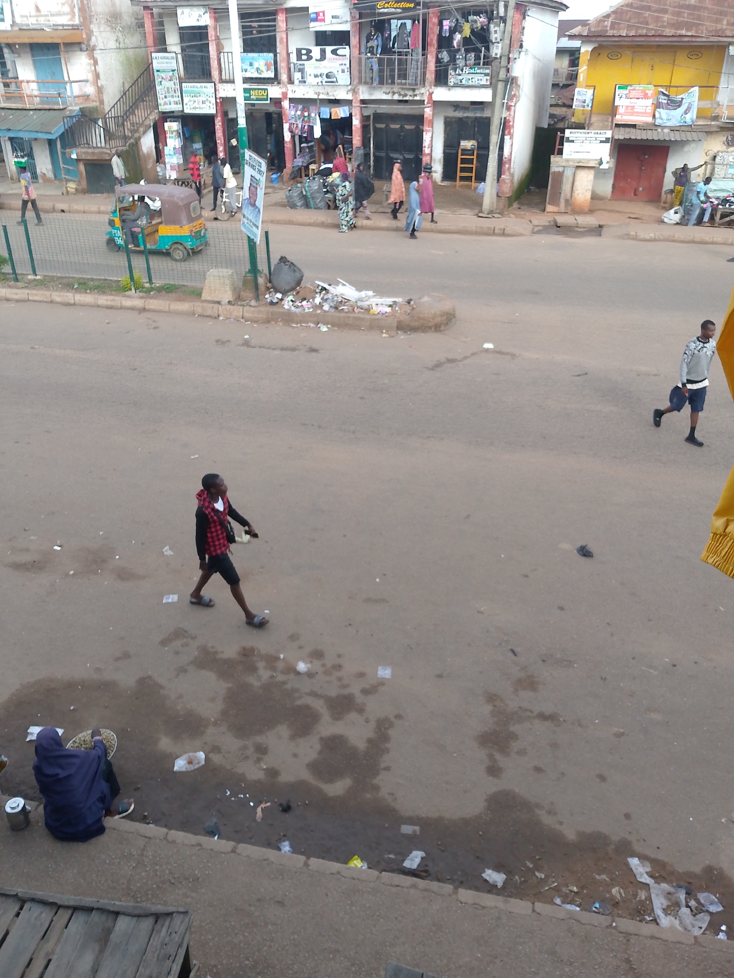 The image depicts a street scene in Bukuru, Nigeria. In the foreground, a person wearing a blue hooded jacket is seated on the curb, holding a bowl of what appears to be food. Beside them is a metal container. The street itself is paved and shows signs of wear and tear, with puddles of water and scattered debris, including paper and plastic waste. In the middle ground, a person in a red and black plaid shirt and black shorts walks across the street, carrying a light-colored bag. Further into the background, there is a yellow and brown auto-rickshaw with two people inside. The street is lined with buildings that house shops, some of which display signage. One building has stairs leading up to a second level where clothing is displayed. Several people are visible walking along the sidewalks and in the street. One person in the far right middle ground wears a patterned long-sleeved shirt and dark shorts, carrying a bag. The environment appears to be an urban commercial area.