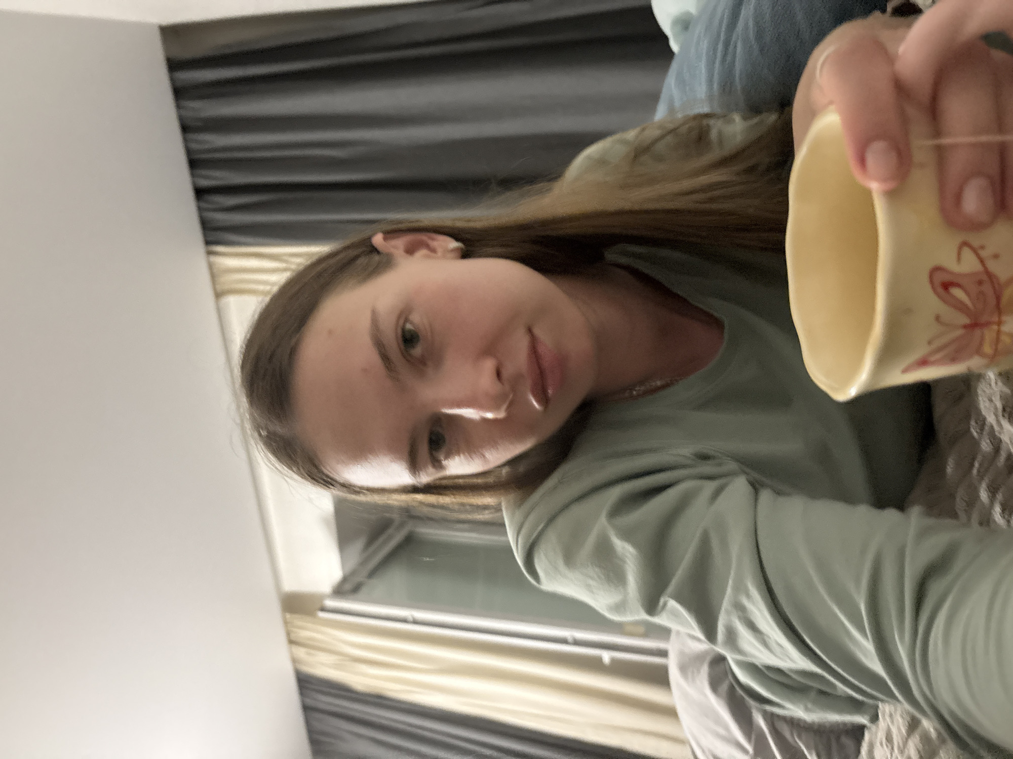 The image shows a young woman lying on a bed, holding a mug of tea. She is wearing a light green long-sleeved shirt. Her hair is long and brown, and she has light-colored eyes. The background consists of curtains and a window, suggesting an indoor setting. The lighting appears to be artificial, possibly from a lamp or overhead light. The scene is casual and relaxed, possibly depicting a morning or evening routine. There are no clear indicators of the specific location within the city of Perm, Russia, or any forensic details that could be extracted.
