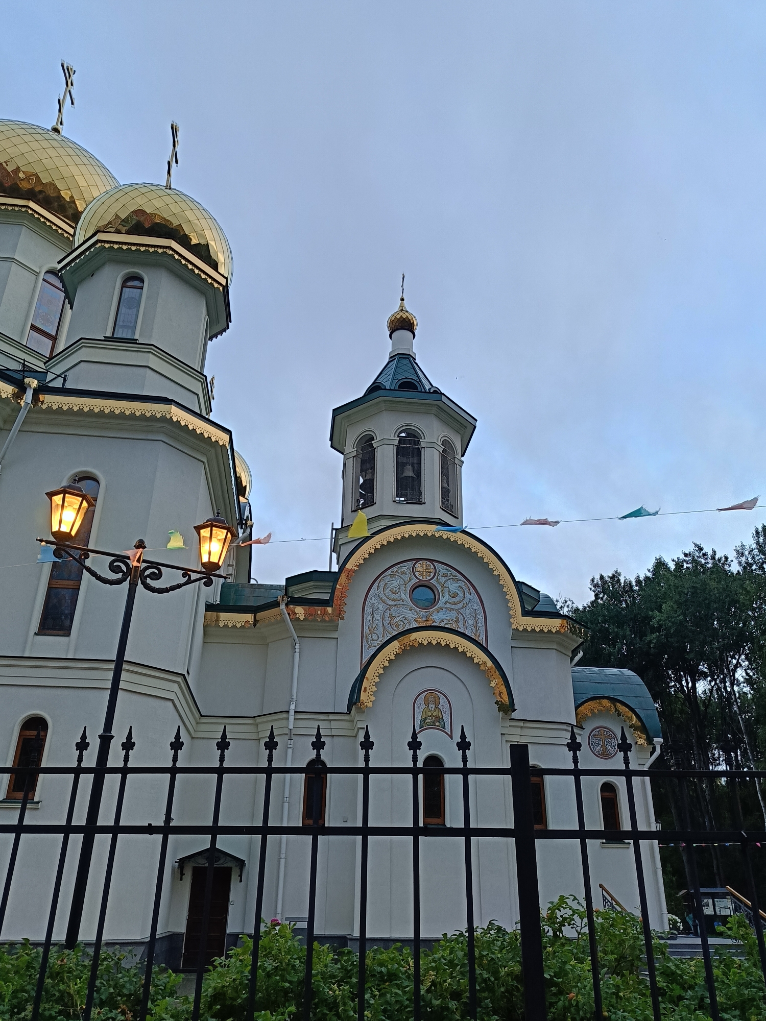 The image depicts the Church of the Icon of the Mother of God Joy of All Who Sorrow in St. Petersburg, Russia, viewed from the outside during overcast daylight. A black wrought-iron fence runs across the foreground, partially obscuring the lower portion of the church and some lush green bushes. Two ornate street lamps with glowing yellow lights are visible on the left side of the frame, mounted on a dark pole with decorative scrolls.
The church itself is predominantly white with gold-domed roofs and intricate gold detailing along its cornices and arched windows. Several onion domes topped with crosses are visible, their golden surfaces reflecting the muted light. One prominent bell tower with a blue roof and a golden dome stands to the right of the main structure. Small, colorful flags or pennants are strung across wires between the street lamps and the church, adding a festive touch to the scene. The sky is a pale, cloudy blue. In the background, a dense line of dark green trees forms a backdrop to the church. The overall impression is one of solemnity mixed with a hint of celebration due to the decorative flags. There are no people visible in the image.